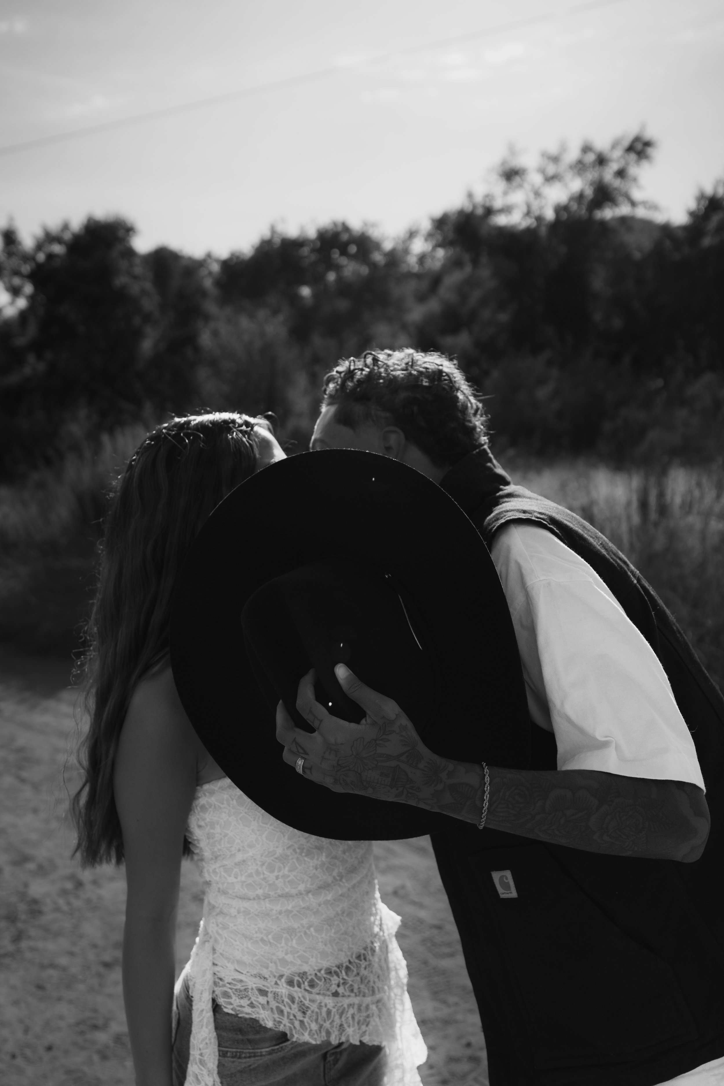 A black and white photo of a couple close together outdoors, with the man holding a wide-brimmed hat near their faces, as they kiss or share an intimate moment.