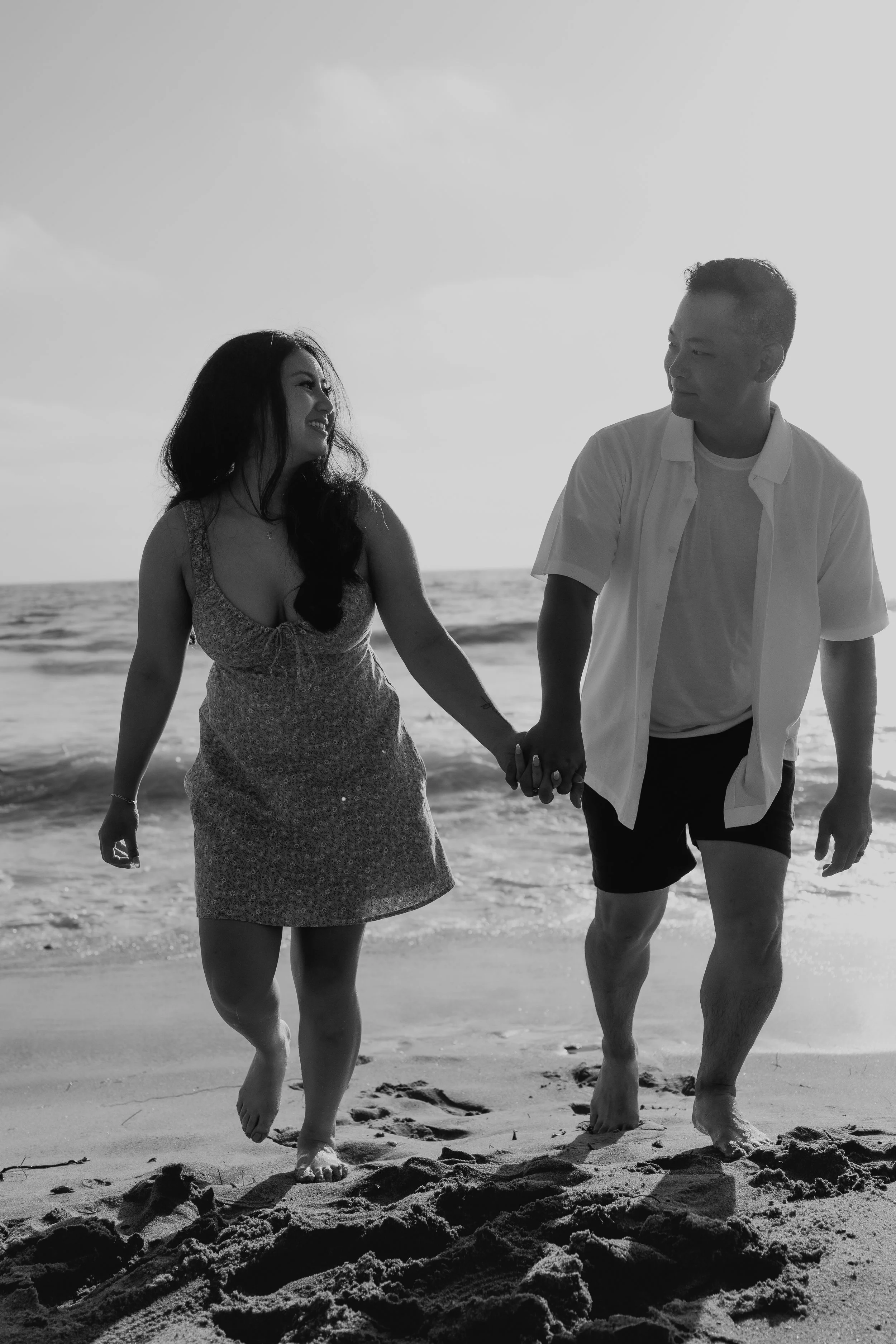 A couple holding hands and walking on the beach, smiling and looking at each other. The woman wears a dress, and the man wears a shirt and shorts. The ocean and sky are in the background.