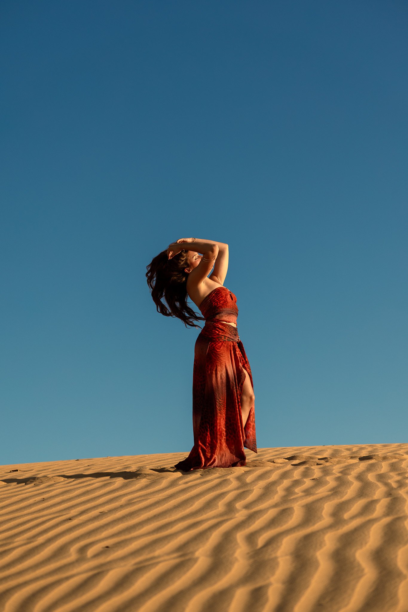 A woman standing on desert sand dunes with blue sky, wearing a red dress, with her arms raised and her hair blowing in the wind.