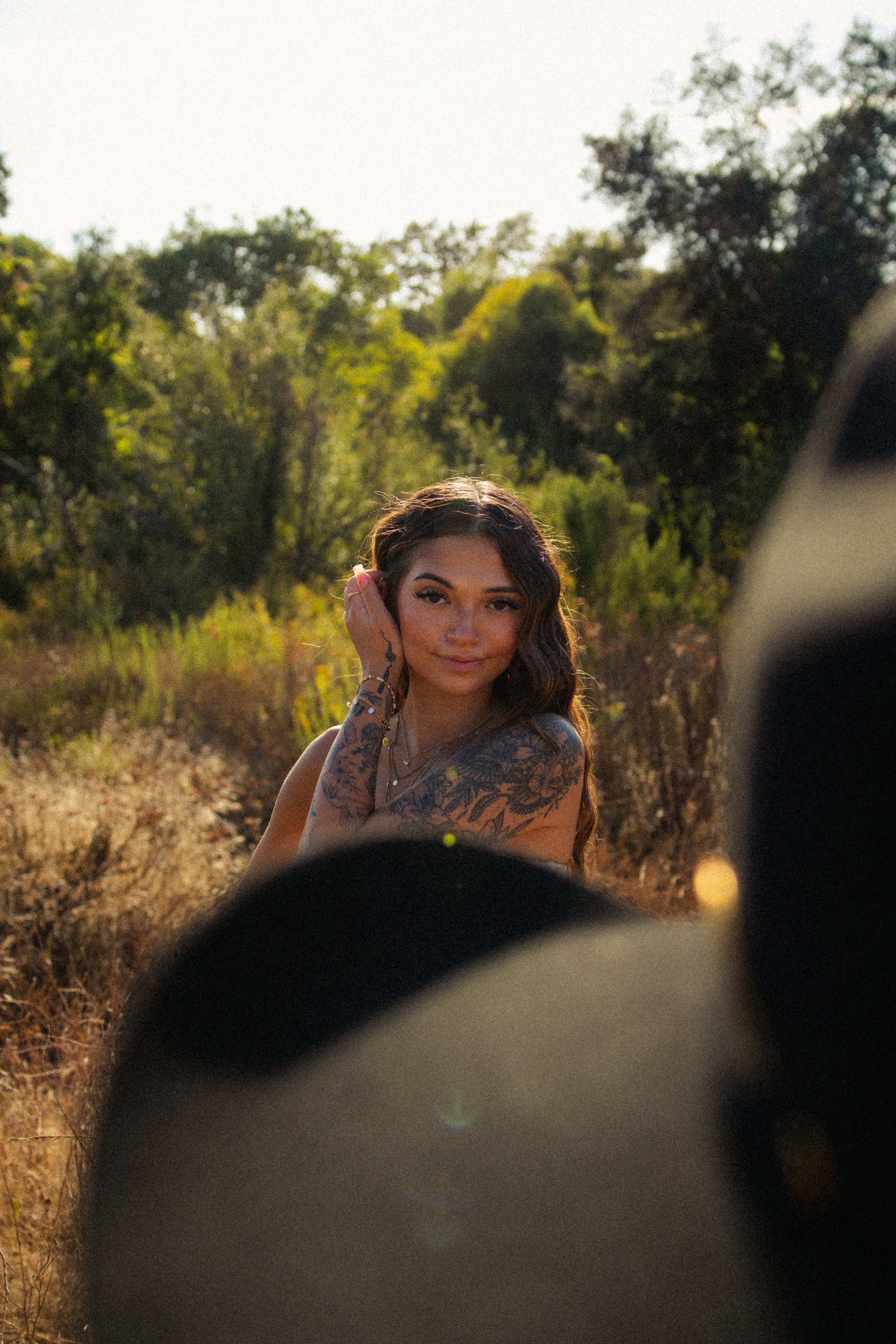 A young woman with long brown hair and tattoos on her arm, smiling and looking at the camera, in a natural outdoor setting with trees and sunlight.