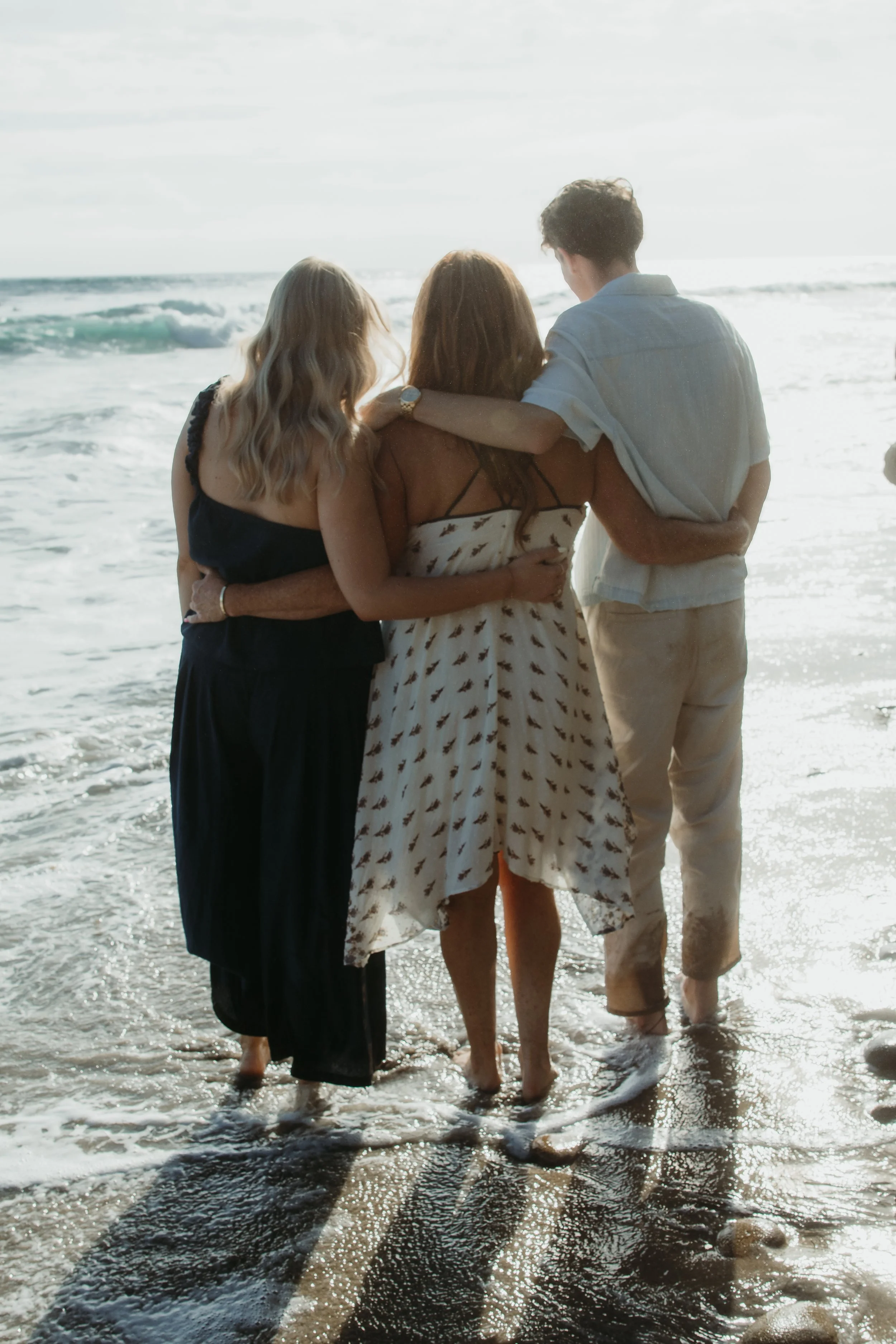 Three people standing close together on the beach with their arms around each other, facing the ocean, in warm sunlight.