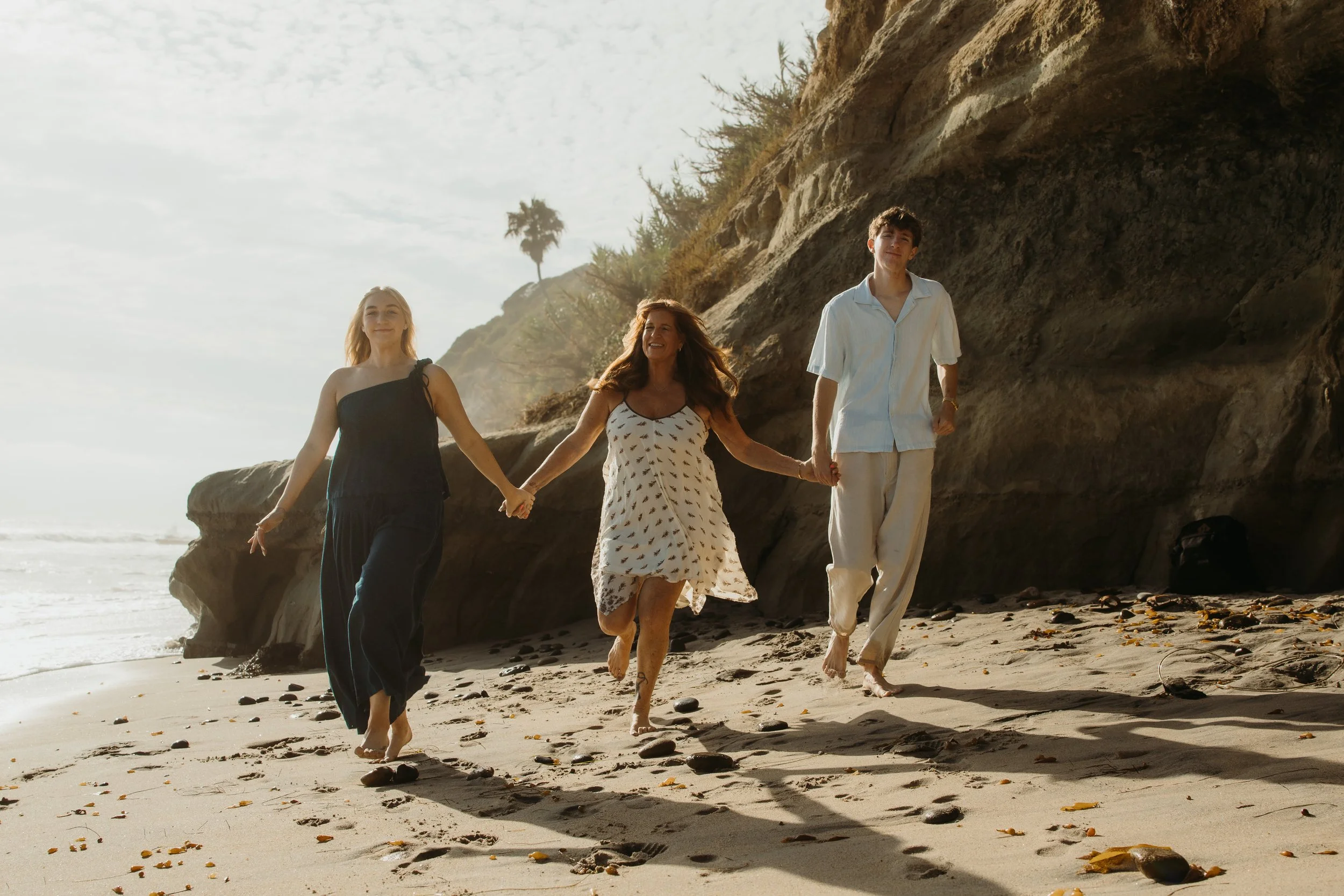 Four people walk hand in hand along the beach near rocks and cliffs, with the ocean and sky in the background.