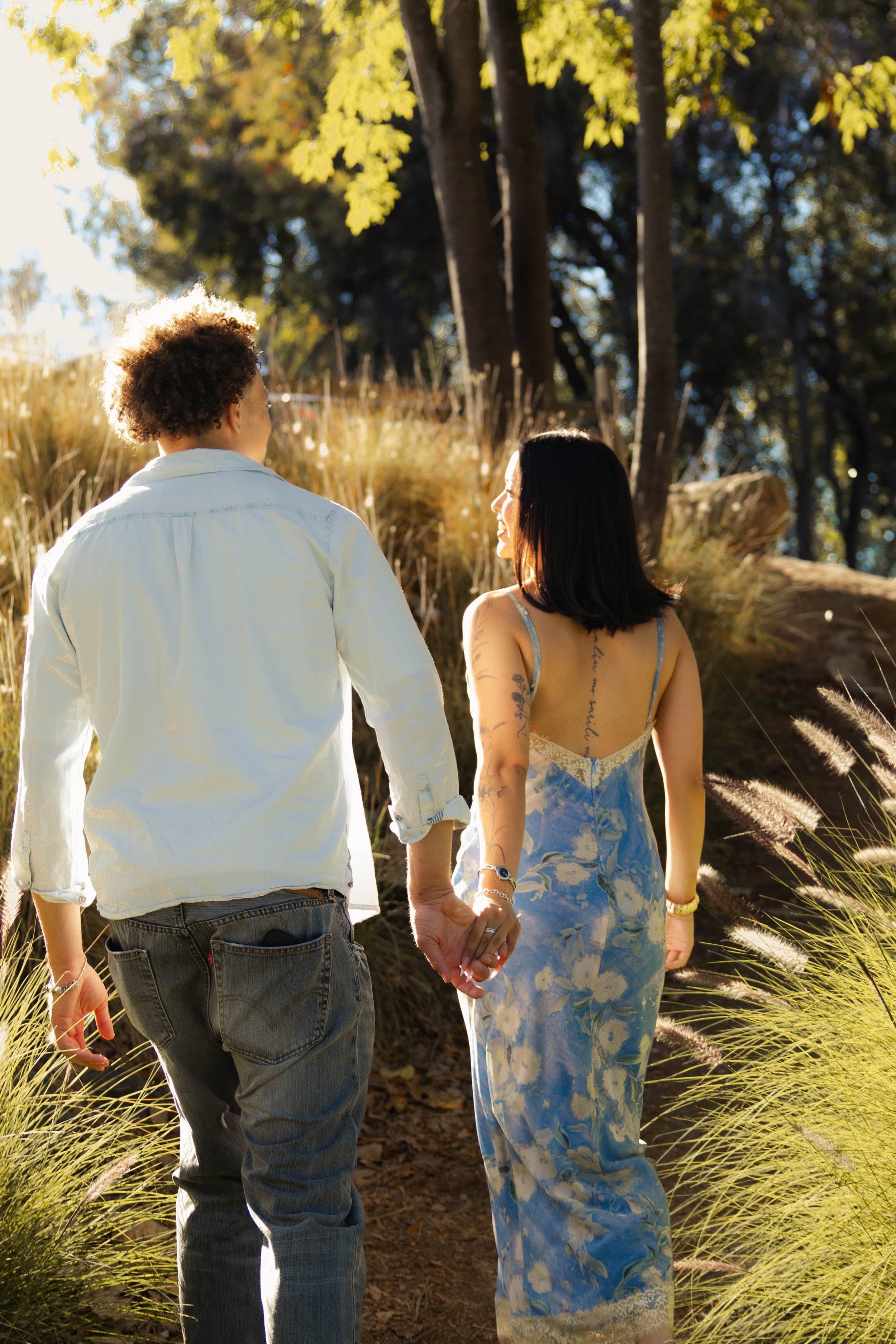 A couple holding hands and walking outdoors on a sunny day, surrounded by trees and tall grass, with the woman wearing a blue floral dress and the man wearing a light shirt and jeans.