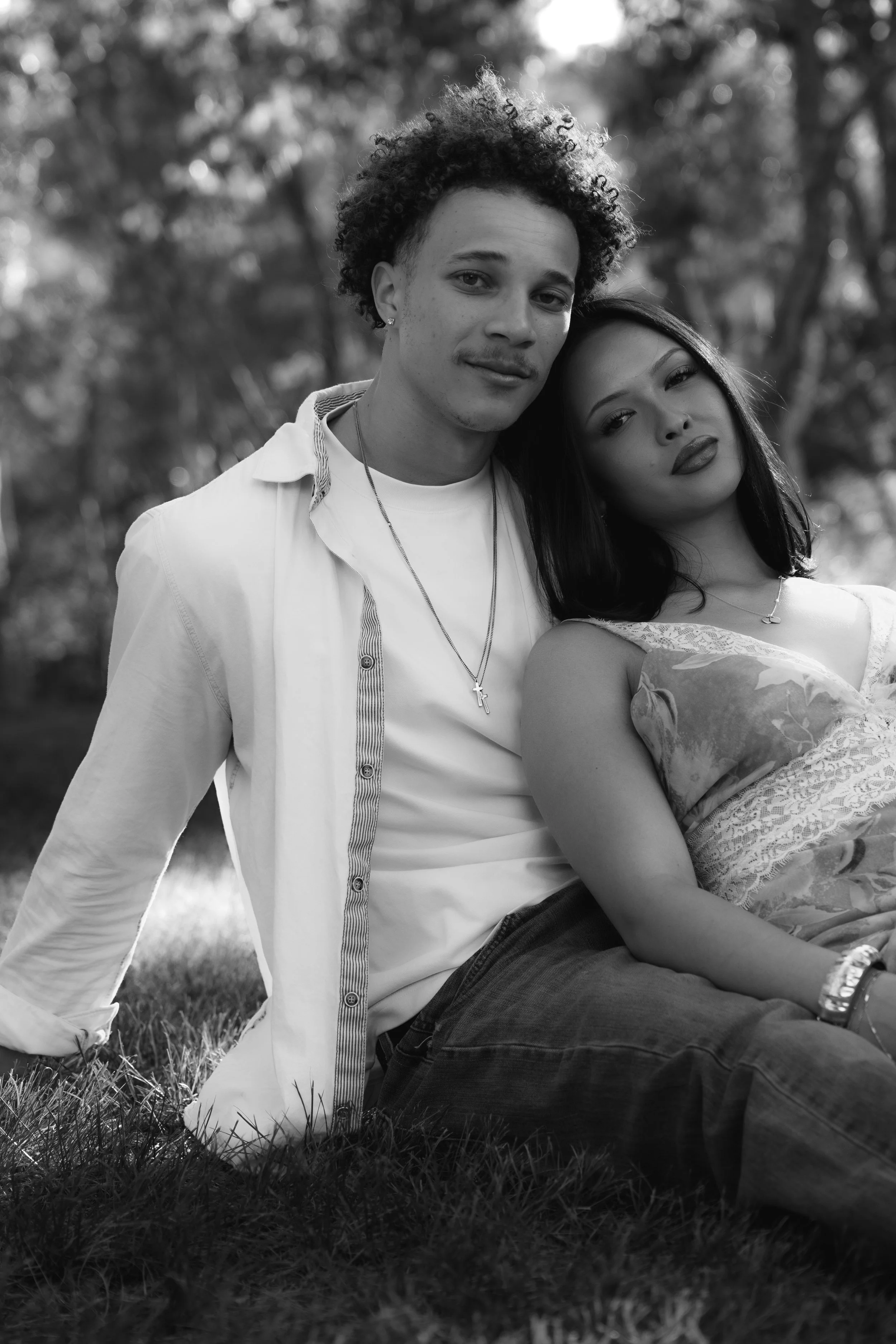 A black and white photograph of a young couple sitting on the grass outdoors, with the man resting his head against the woman's shoulder, both looking at the camera.