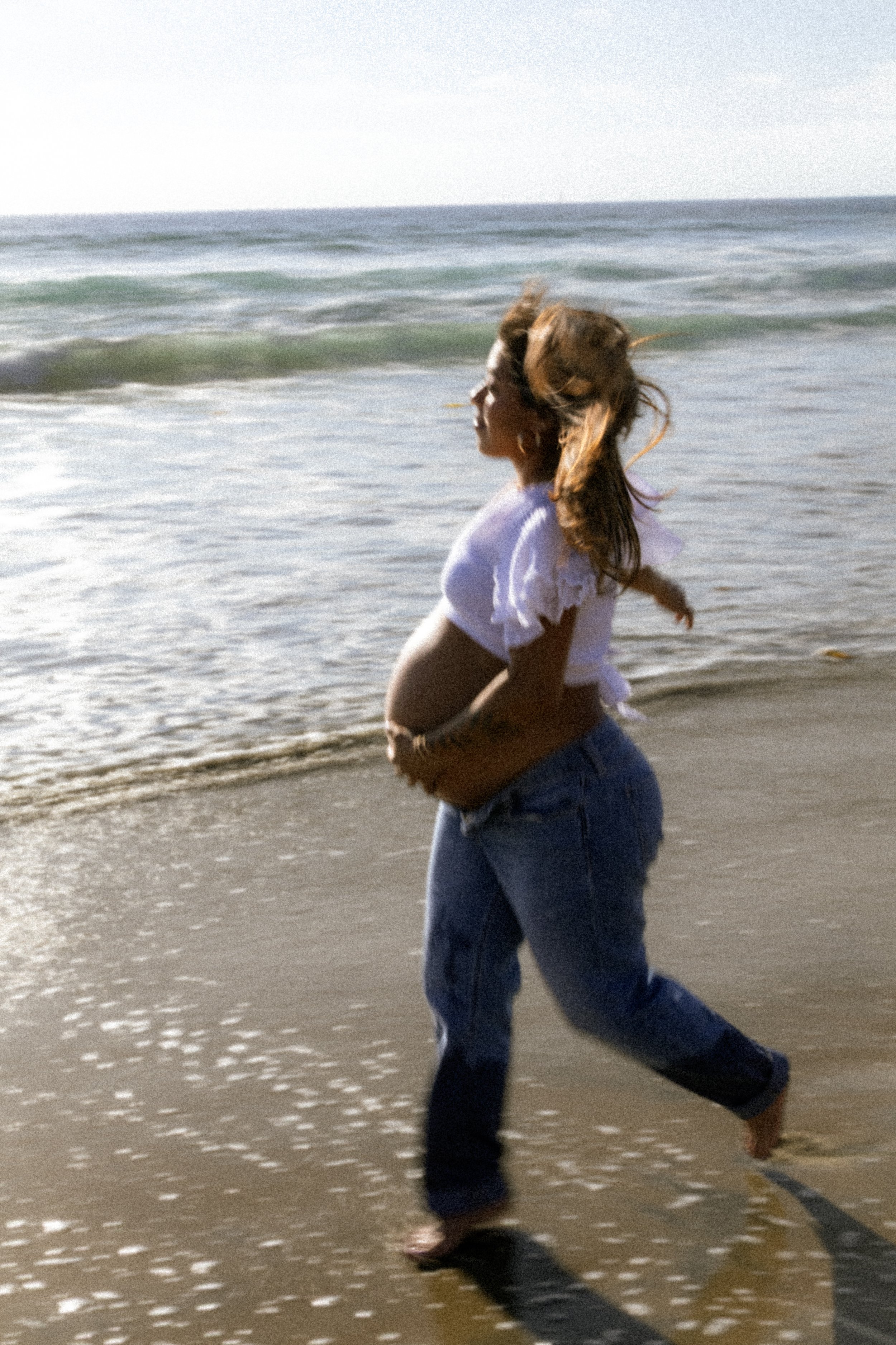 A woman running along the shoreline of a beach with the ocean in the background. She is wearing a white top and blue jeans, with her hair flowing in the wind.