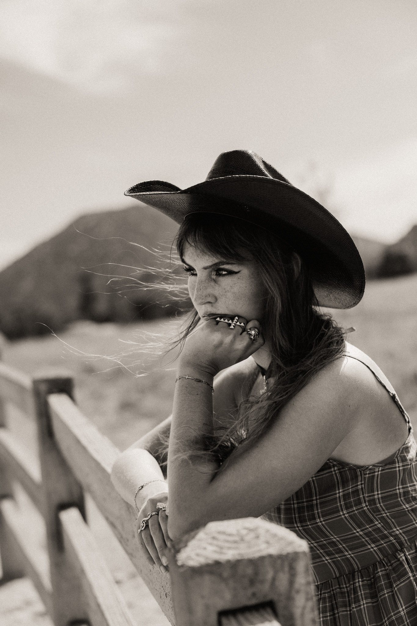 A young woman with long wavy hair, wearing a large wide-brimmed hat and a plaid dress, leaning on a wooden fence on a hill, gazing into the distance on a cloudy day.
