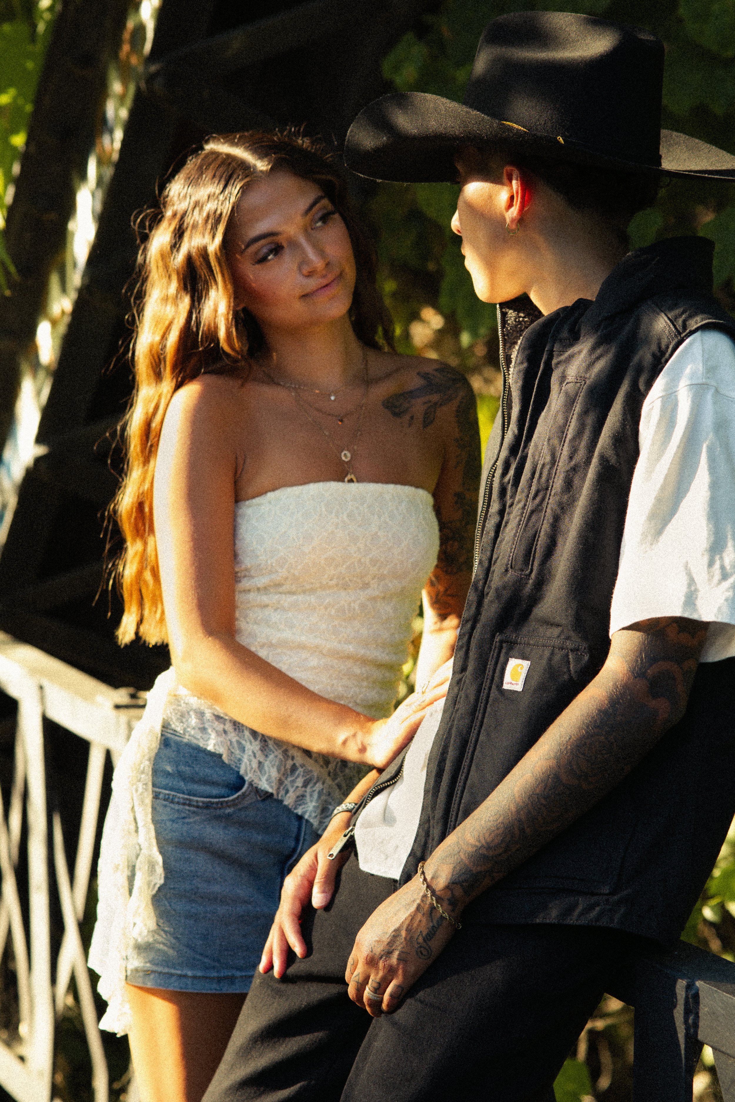 A woman with long, wavy hair and tattoos on her arm looks at a man wearing a black hat and vest.