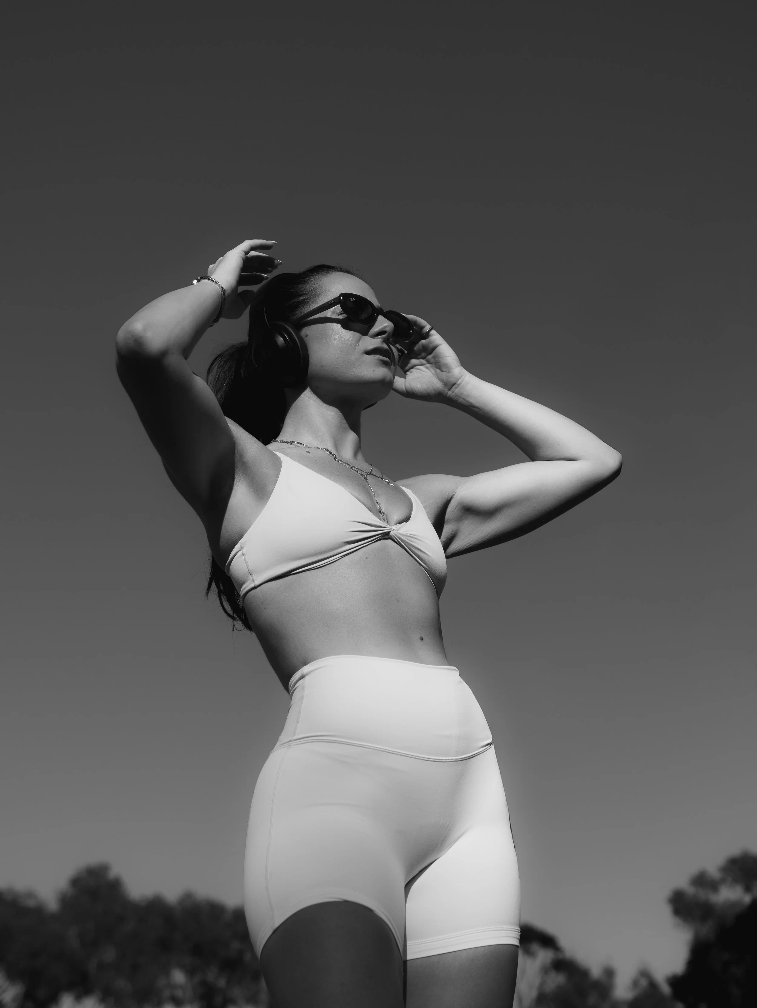 A woman wearing sunglasses and headphones, wearing a tank top and shorts, standing outdoors with a clear sky and some trees in the background.