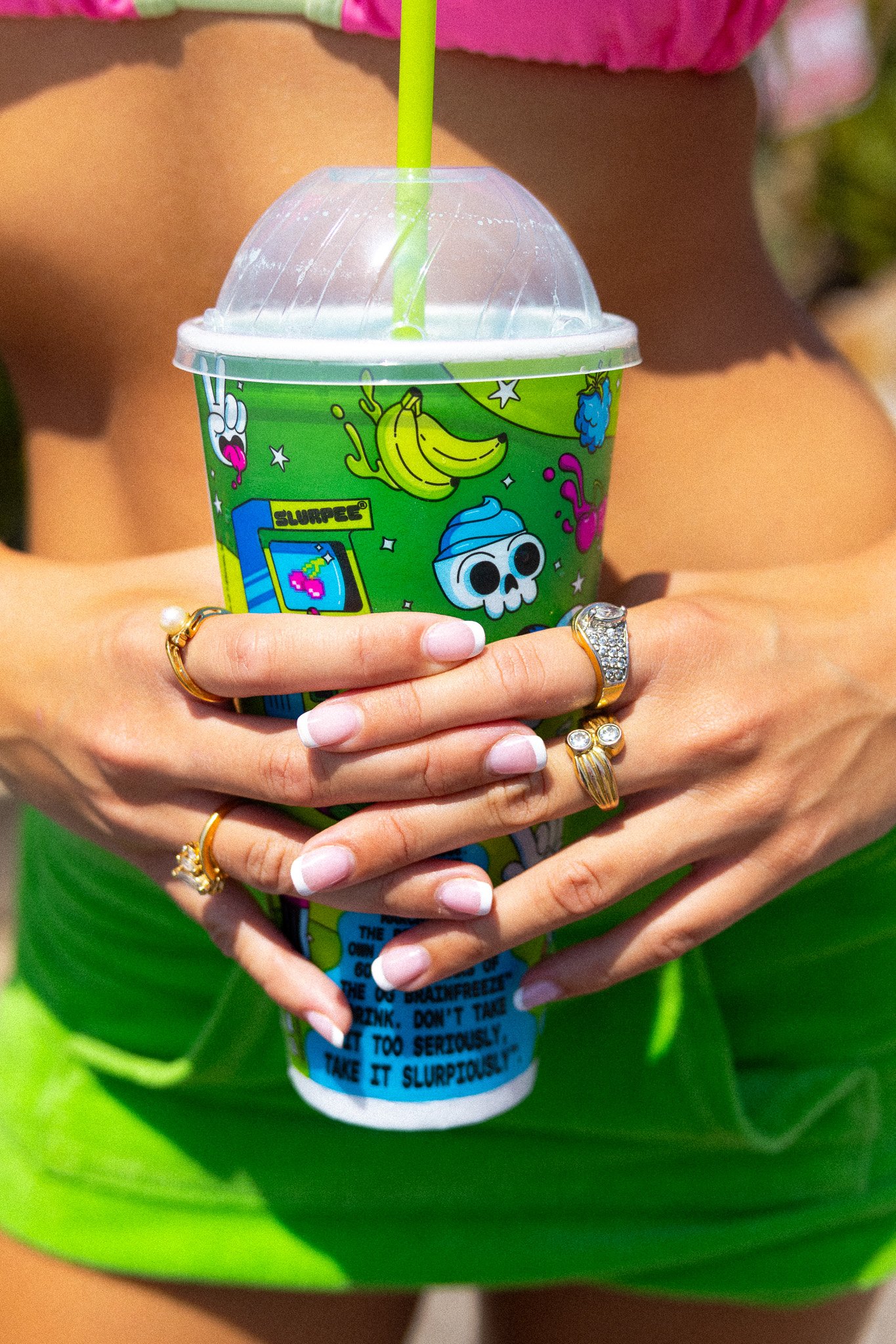 Close-up of a person holding a colorful slush cup with a green straw. The person is wearing multiple rings on fingers with French manicured nails, and a green dress with pink top bottom. The cup has vibrant graphics including a skull, bananas, and va