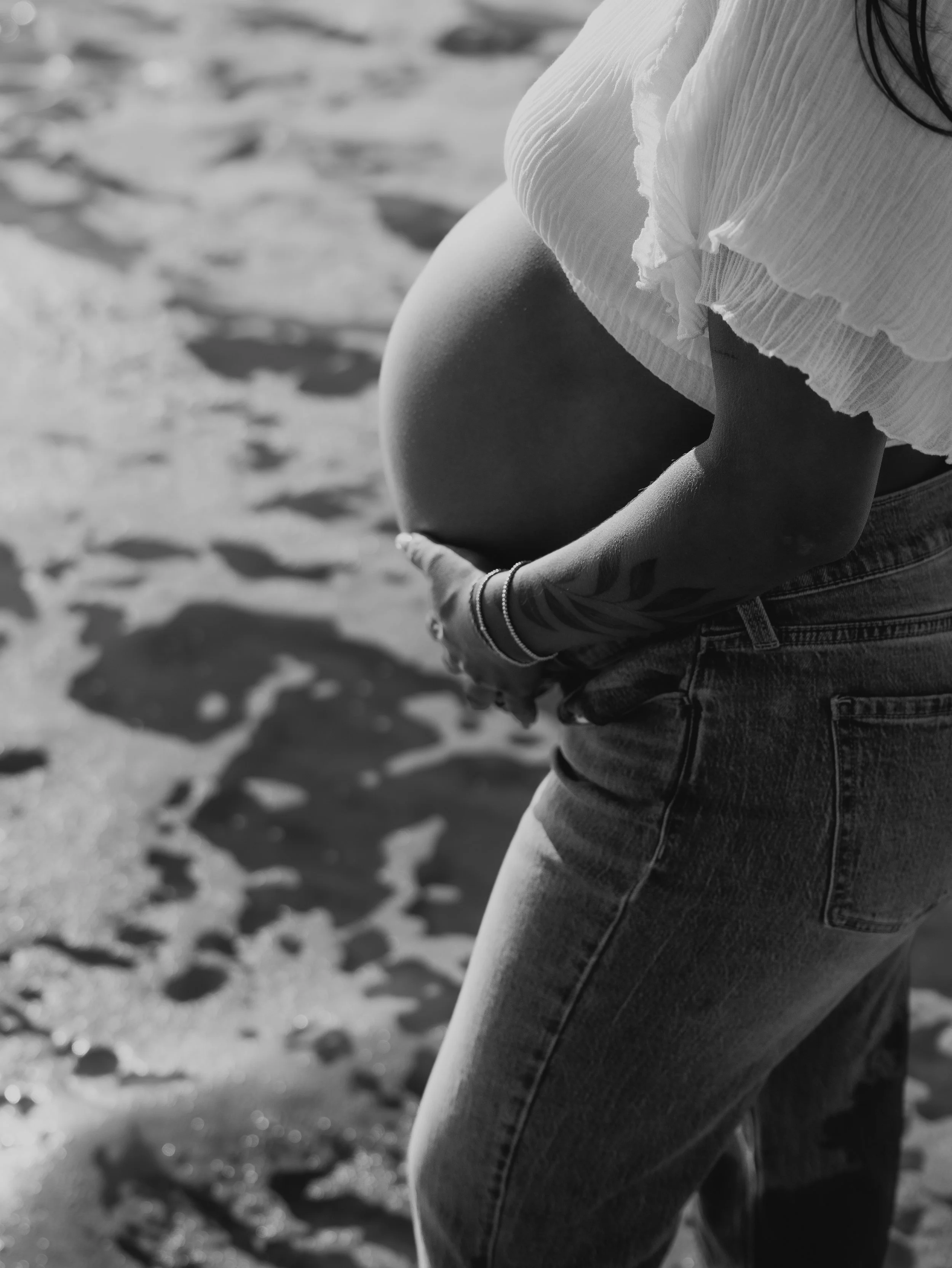 Close-up of a woman's pregnant belly covered by a flowing white dress, standing on a sandy beach.