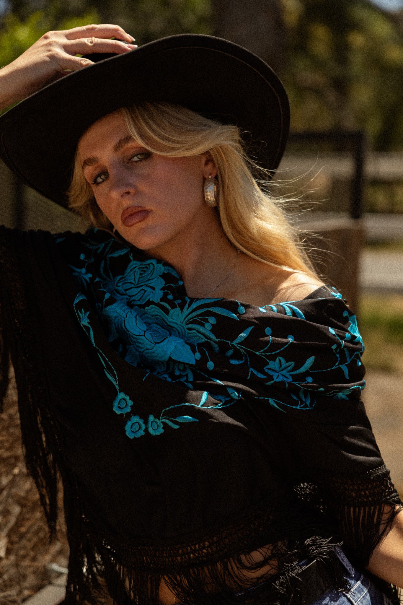 A woman with blonde hair wearing a black wide-brimmed hat, a black garment with blue floral embroidery, and silver hoop earrings. She is sitting outdoors with a blurred background of greenery.