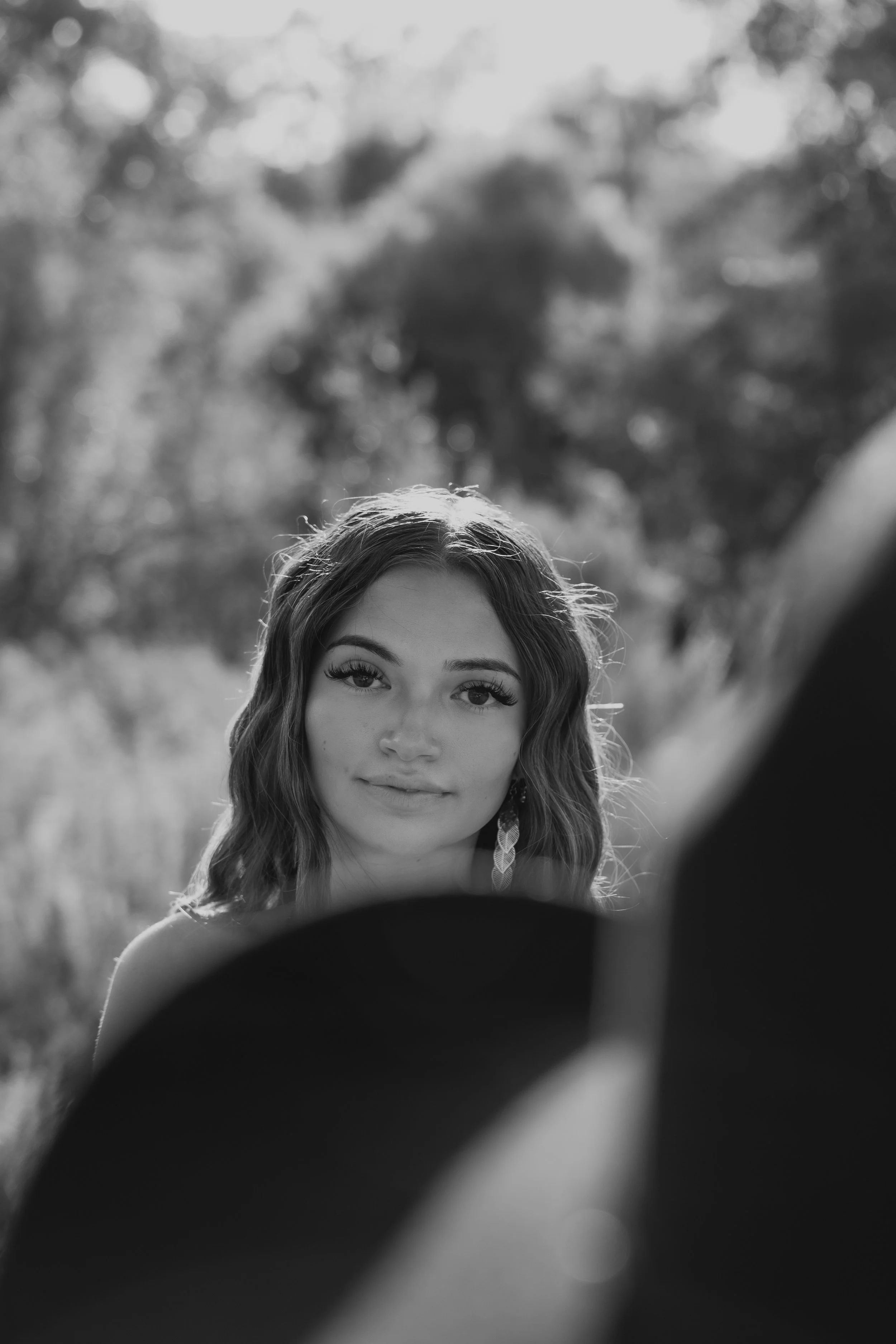 Black and white photo of a woman with long wavy hair, looking into a camera, outdoors with blurred trees in the background, and an out-of-focus object in the foreground.