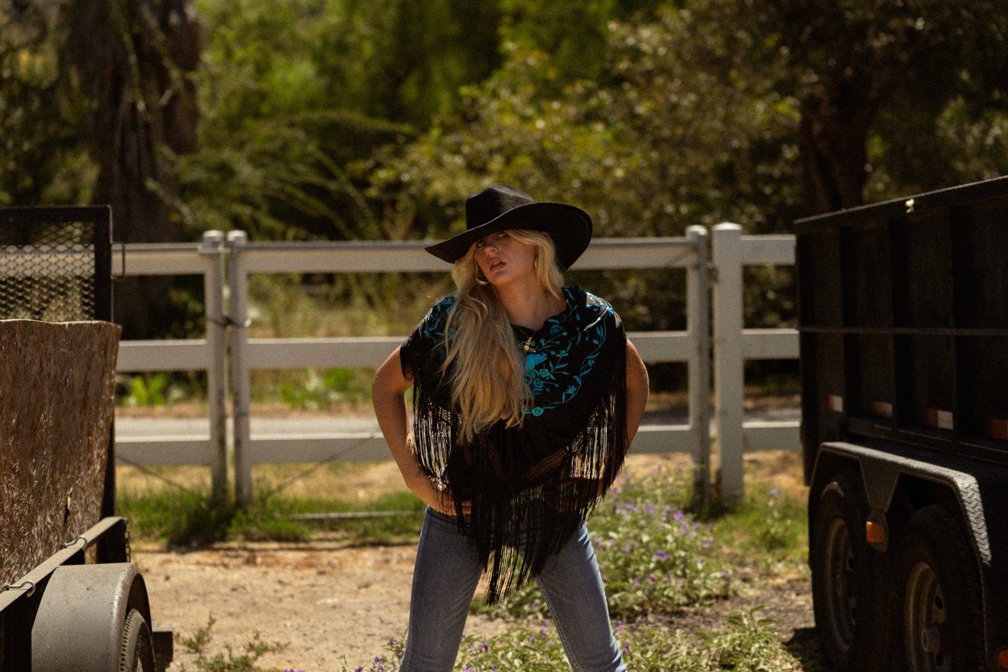A woman with long blonde hair wearing a wide-brimmed black hat, a black top with blue floral designs, and fringed shawl, standing outdoors near a fence and a black vehicle, with trees and greenery in the background.