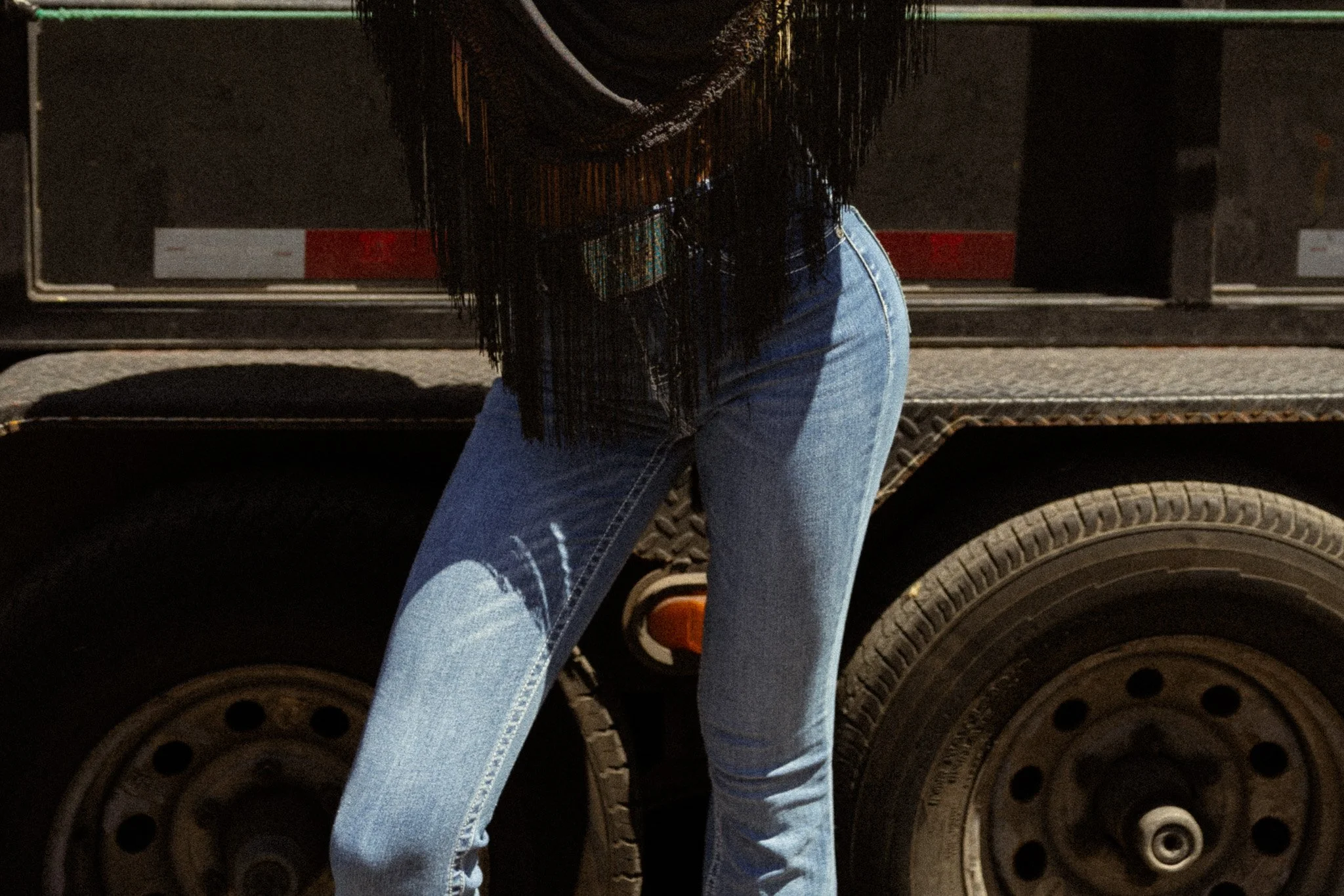 Part of a person wearing a black shirt and cut-off blue jeans, standing in front of a black truck with large tires, digital photo taken outdoors at night.