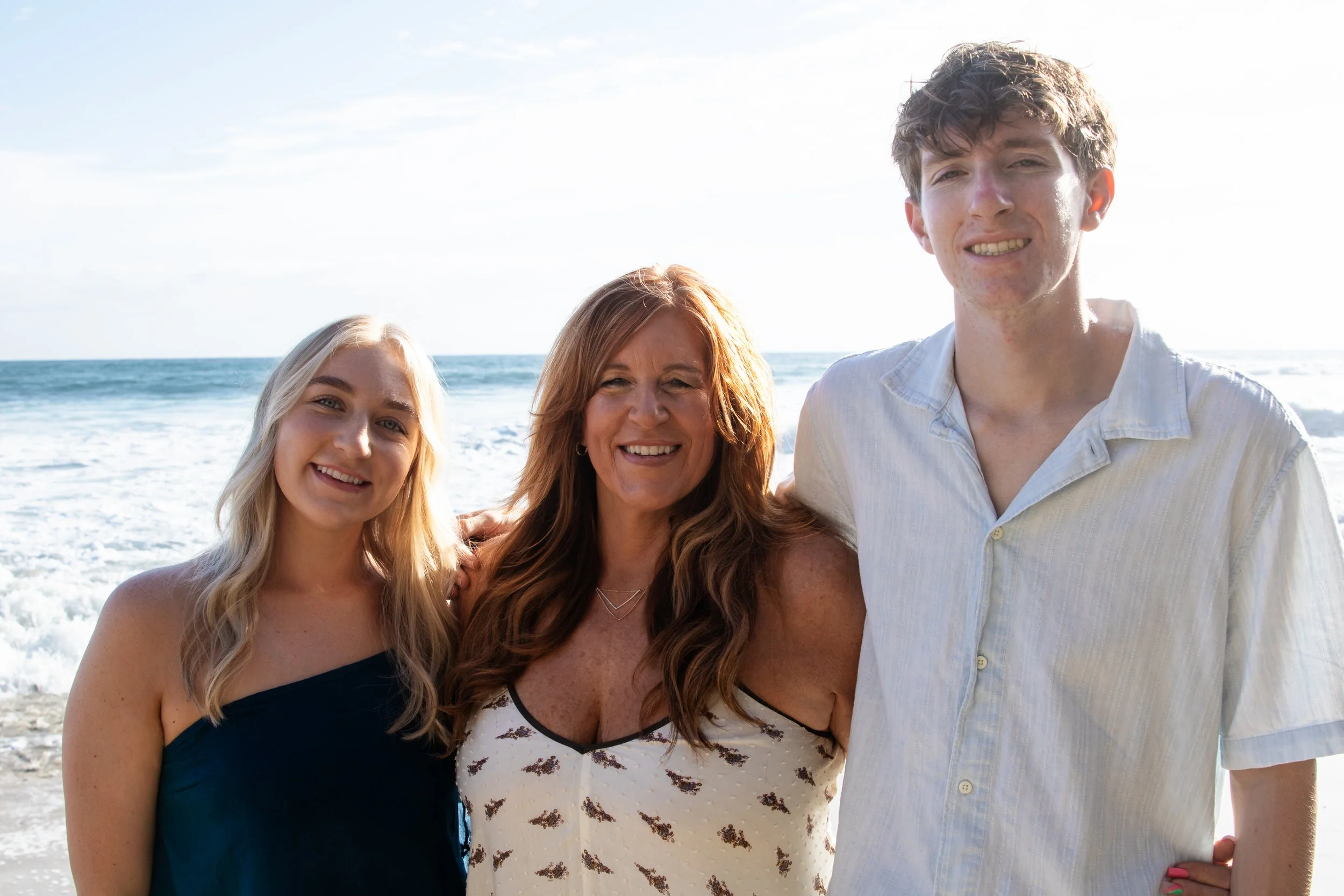 A smiling woman with long brown hair standing between a teenage girl with long blonde hair and a young man with curly brown hair, at the beach with the ocean in the background.