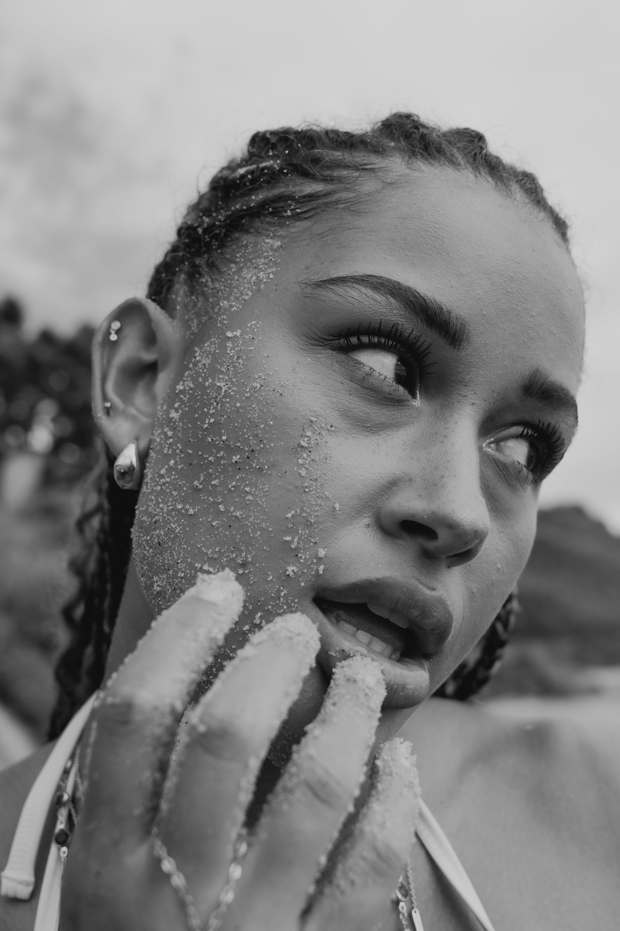 Close-up black and white photo of a young woman with a radiant smile, showing her teeth. She has curly hair tied back and is wearing earrings and layered necklaces. She is holding a piece of chalk near her face with a background of an overcast sky.