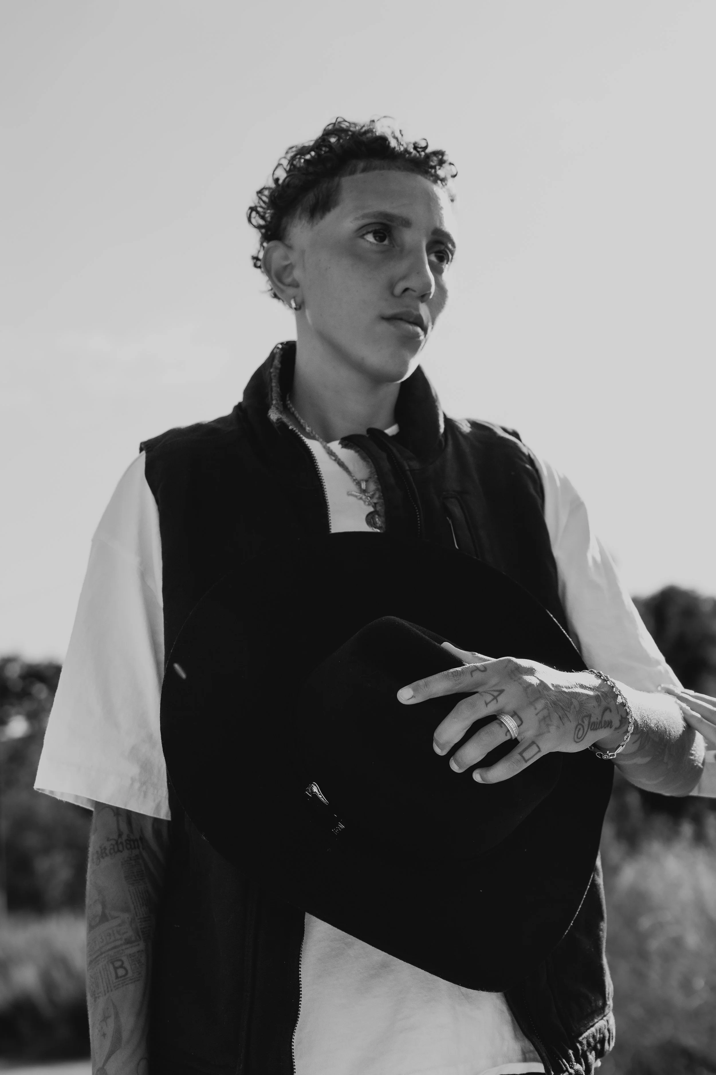 A black-and-white photo of a young person with short curly hair, wearing earrings, a layered necklace, and a dark vest over a light T-shirt. They are holding a black helmet with one hand and have tattoos on their arm, with tattoos and rings on their 