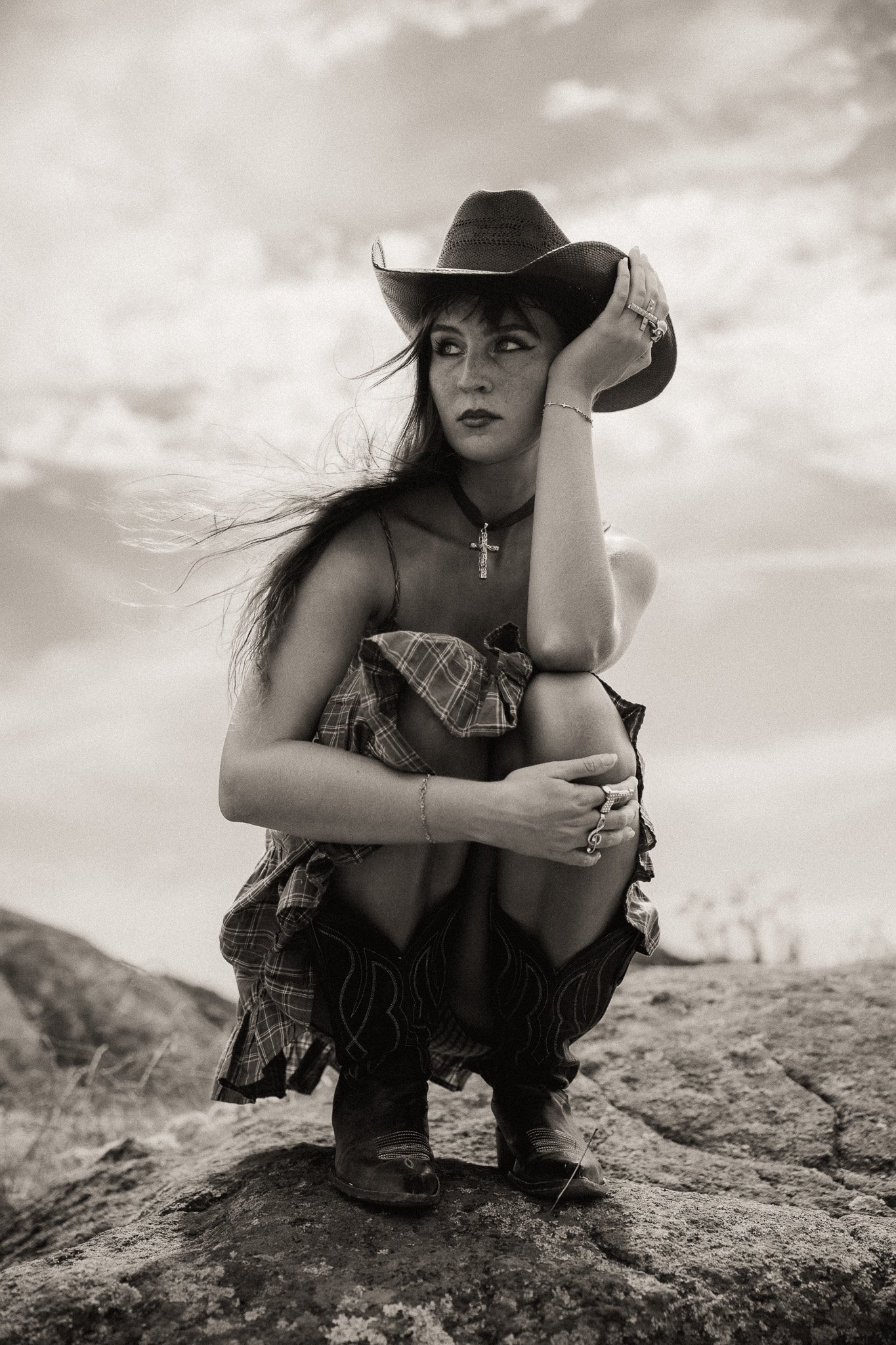 A woman with long hair wearing a cowboy hat and jewelry, crouching on a rock outdoors. She is dressed in a plaid shirt tied around her waist, a dark top, patterned pants, and boots. She is looking away with her hand on her head and arm crossed over h