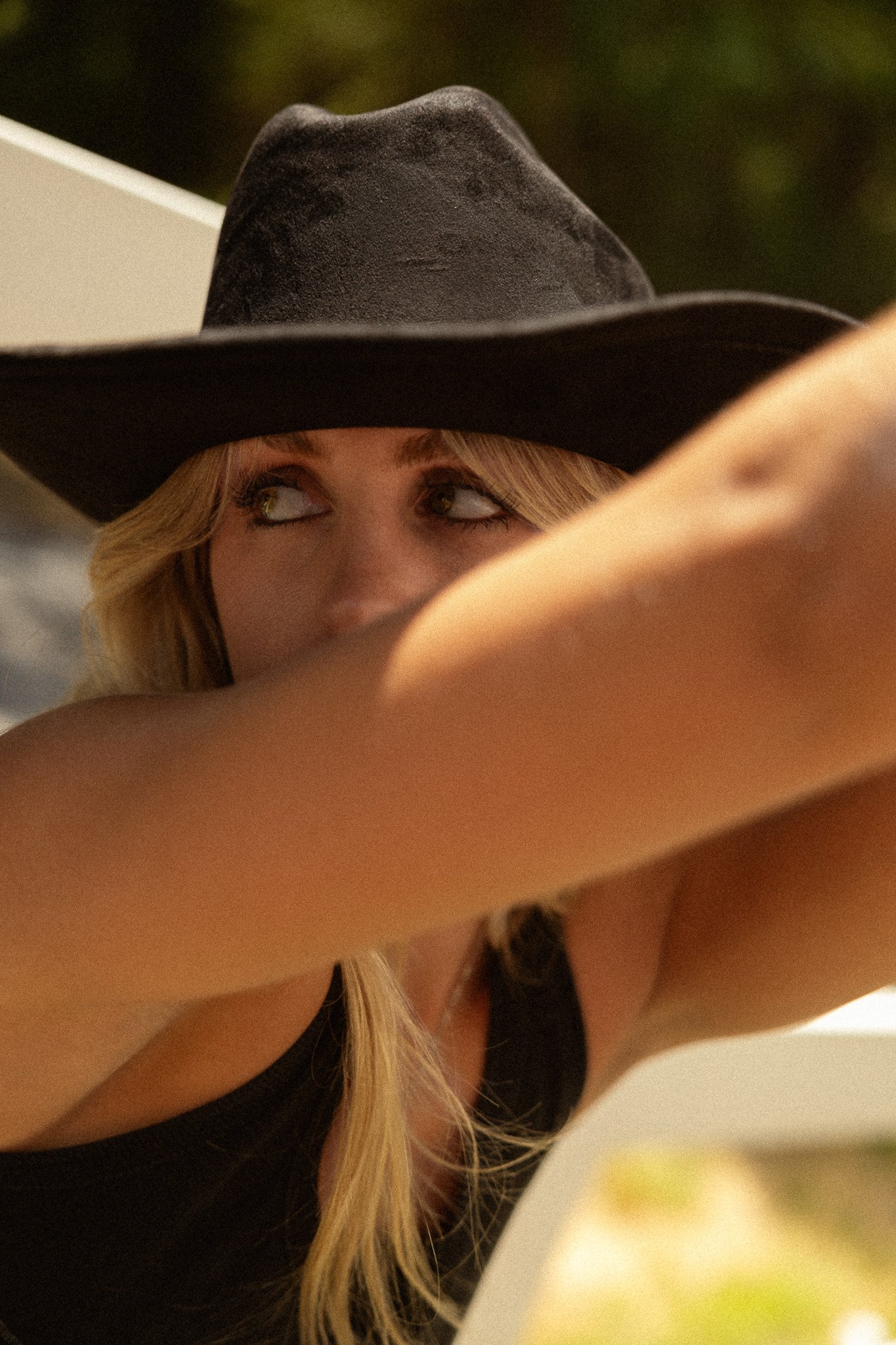 A woman with long blonde hair wearing a black wide-brimmed hat and a black top, looking to her left with a blurred background.
