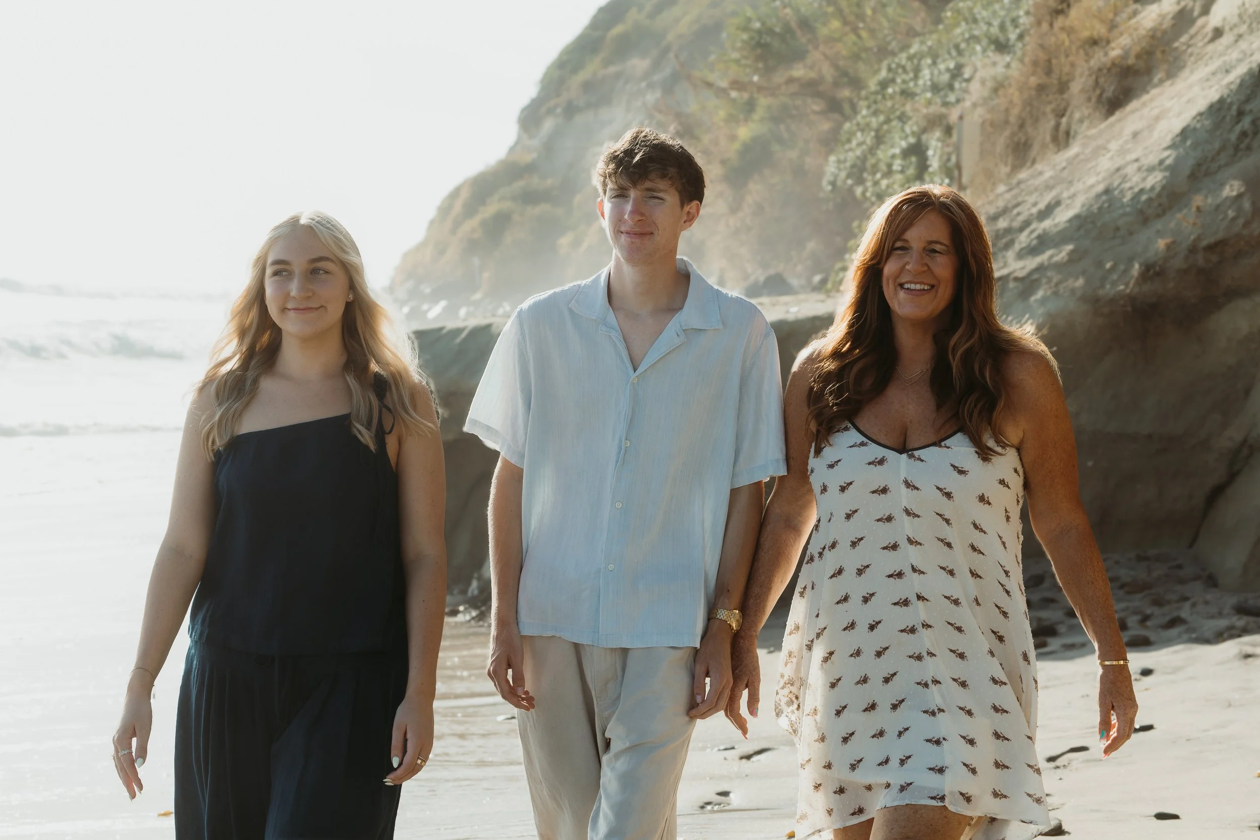 Three women and one man walking along the beach near rocky cliffs, with ocean waves in the background on a sunny day.