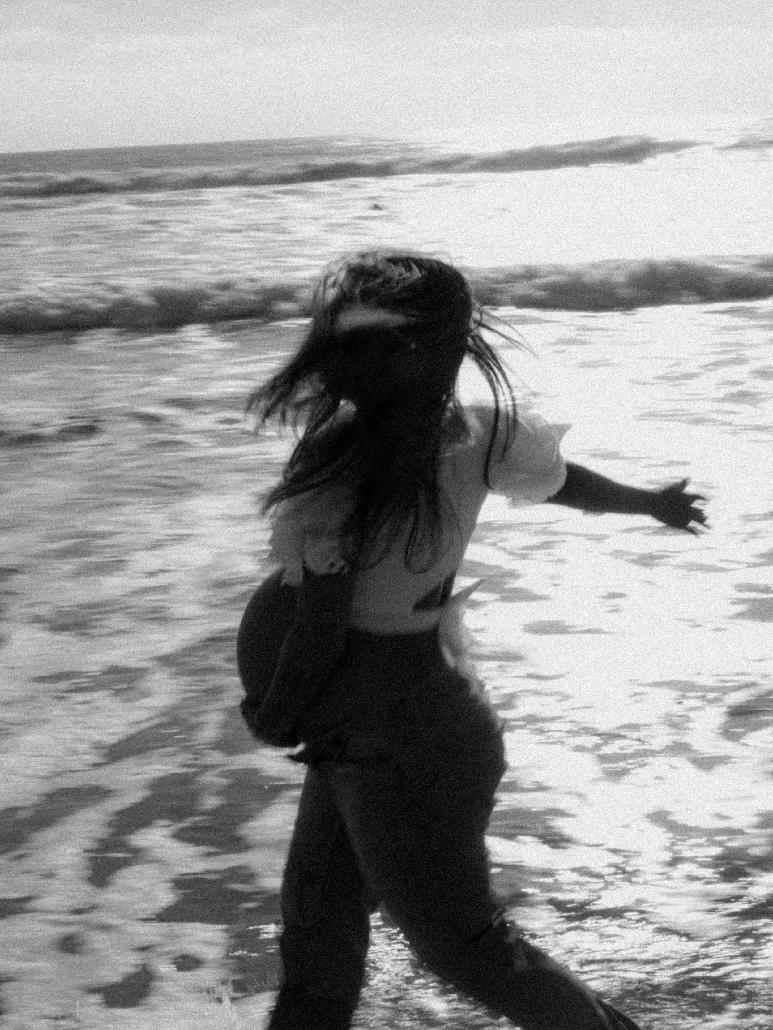 A woman with long hair standing on the beach with waves in the background, in black and white.