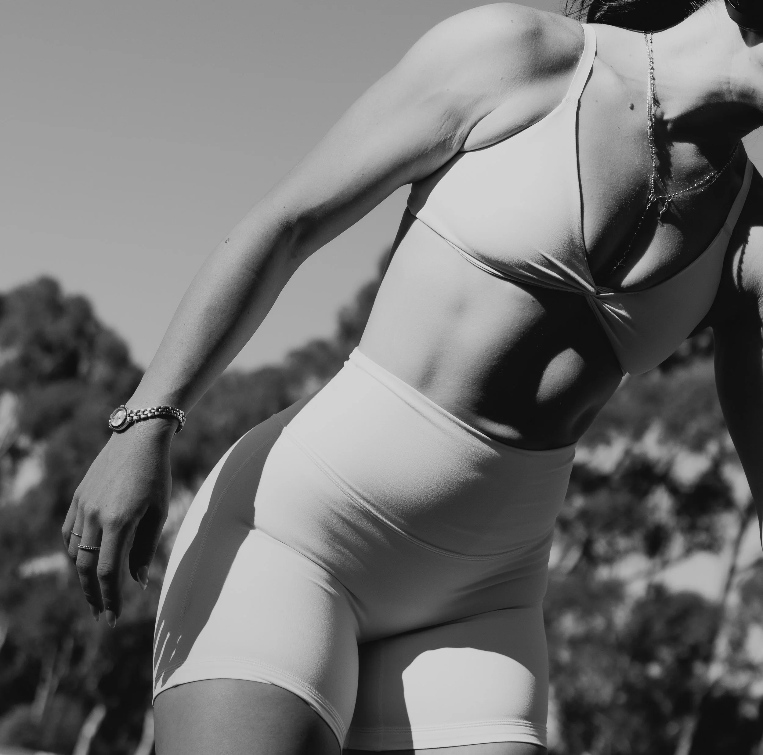 A woman in a sports bra and shorts stretching outdoors on a sunny day, wearing a bracelet and a necklace.
