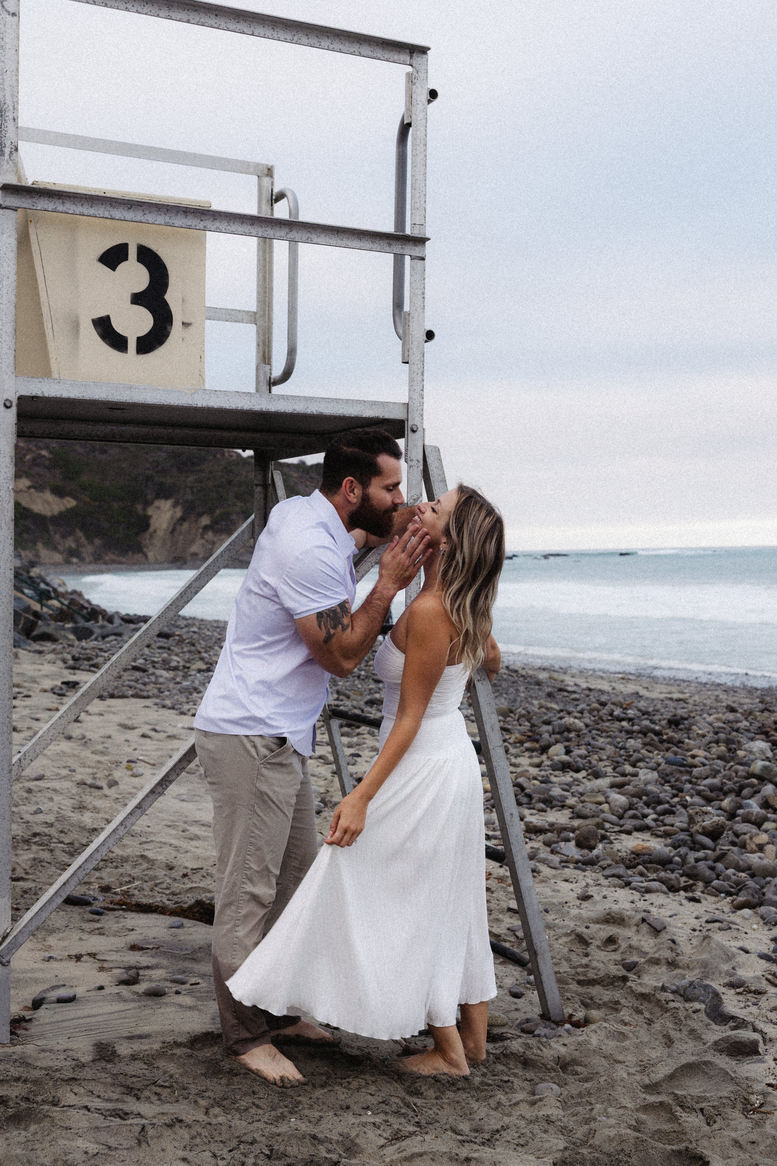 A couple stands on a pebble and sand beach, holding hands and gazing into each other's eyes, near a lifeguard tower with the number 3, overlooking the ocean.