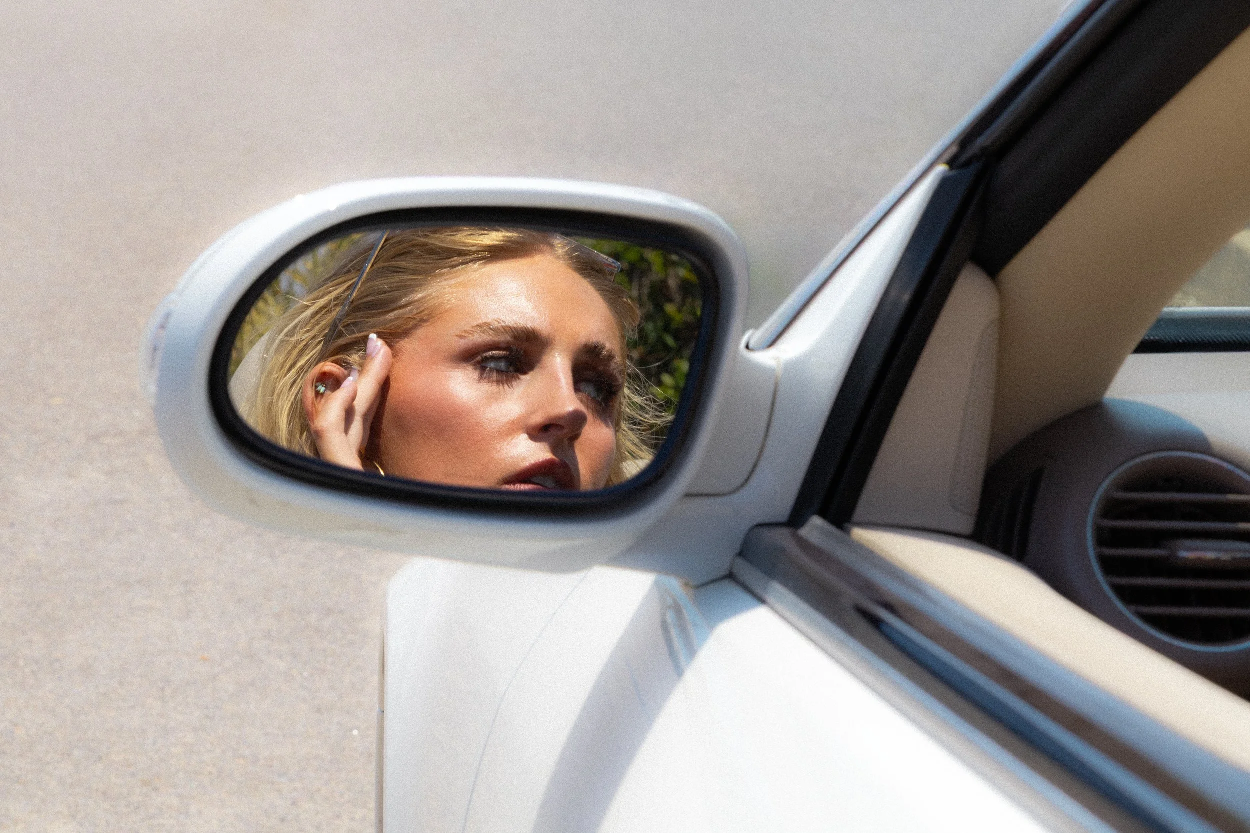 Close-up of a woman with blonde hair in the side mirror of a white car, touching her temple with her hand and looking at the camera.
