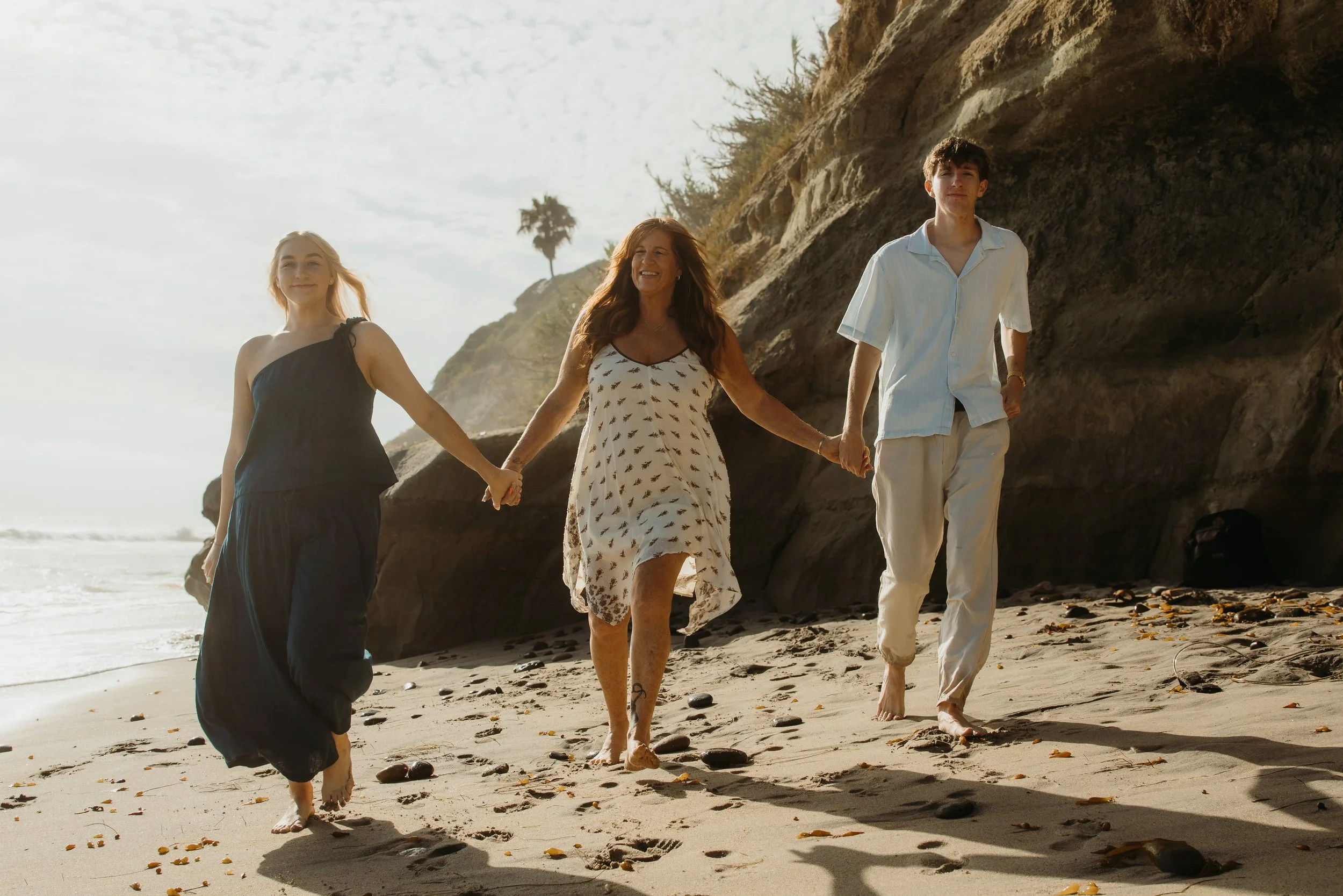 Three people, one older woman and two younger individuals, holding hands and walking barefoot on a sandy beach with rocks and a large cliff in the background.