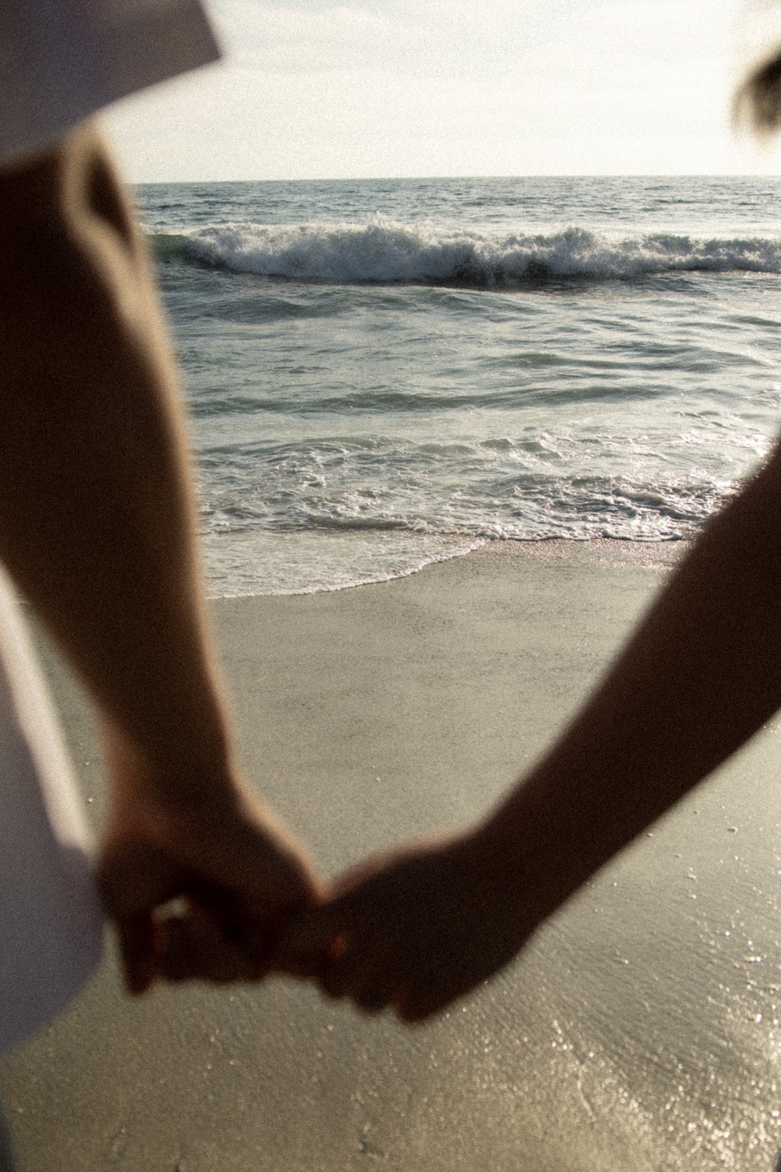 Photo of the beach, framed by the hands of a person making a heart shape, with the ocean waves in the background.