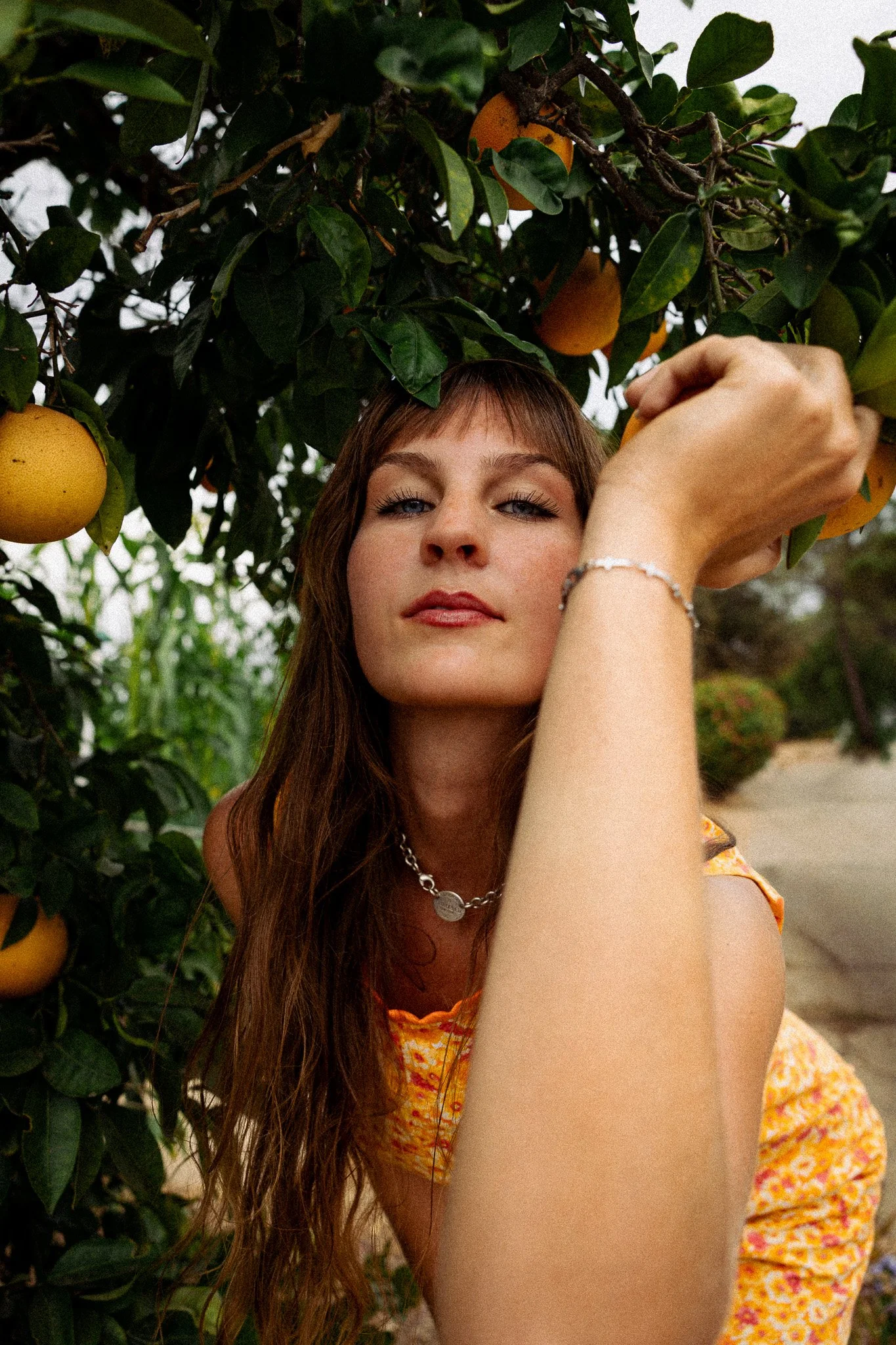 A woman with long brown hair and blue eyes is standing outdoors among green foliage with yellow oranges, partially obscuring her face. She's wearing a yellow patterned dress, a silver bracelet, and a necklace.