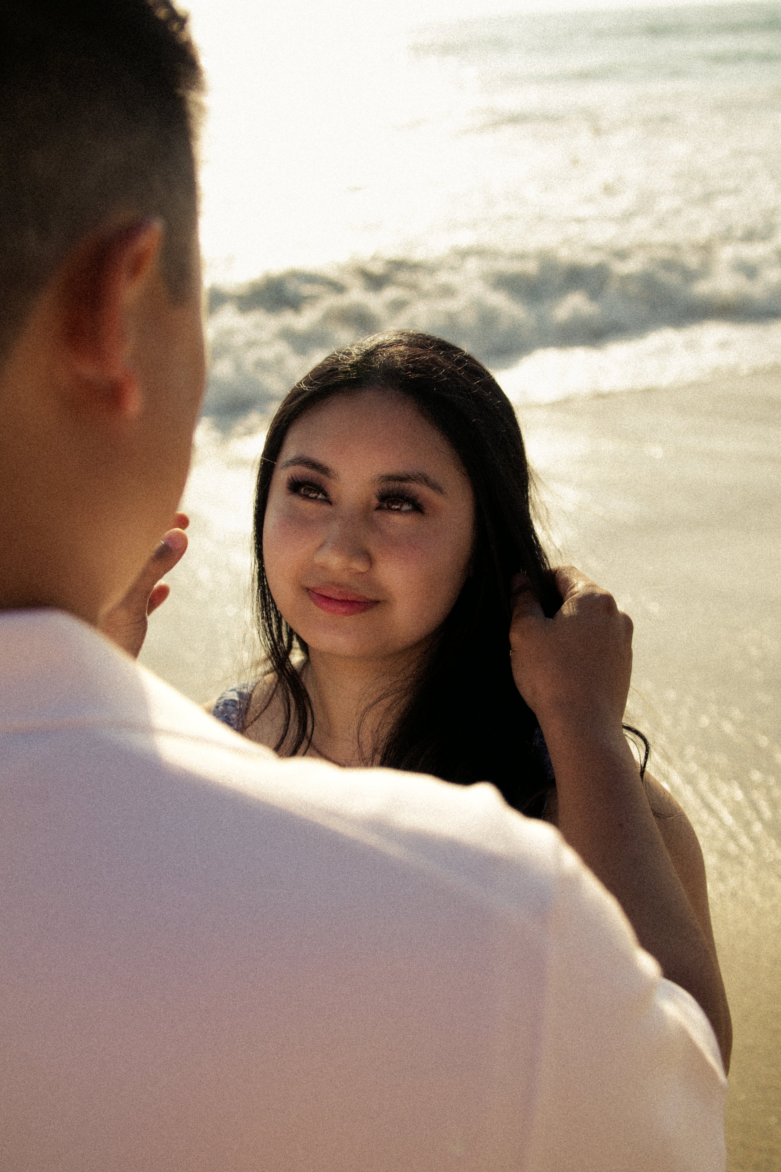 A man and woman on the beach. The woman is smiling, looking up at the man, with the ocean waves in the background, early morning or late afternoon lighting.