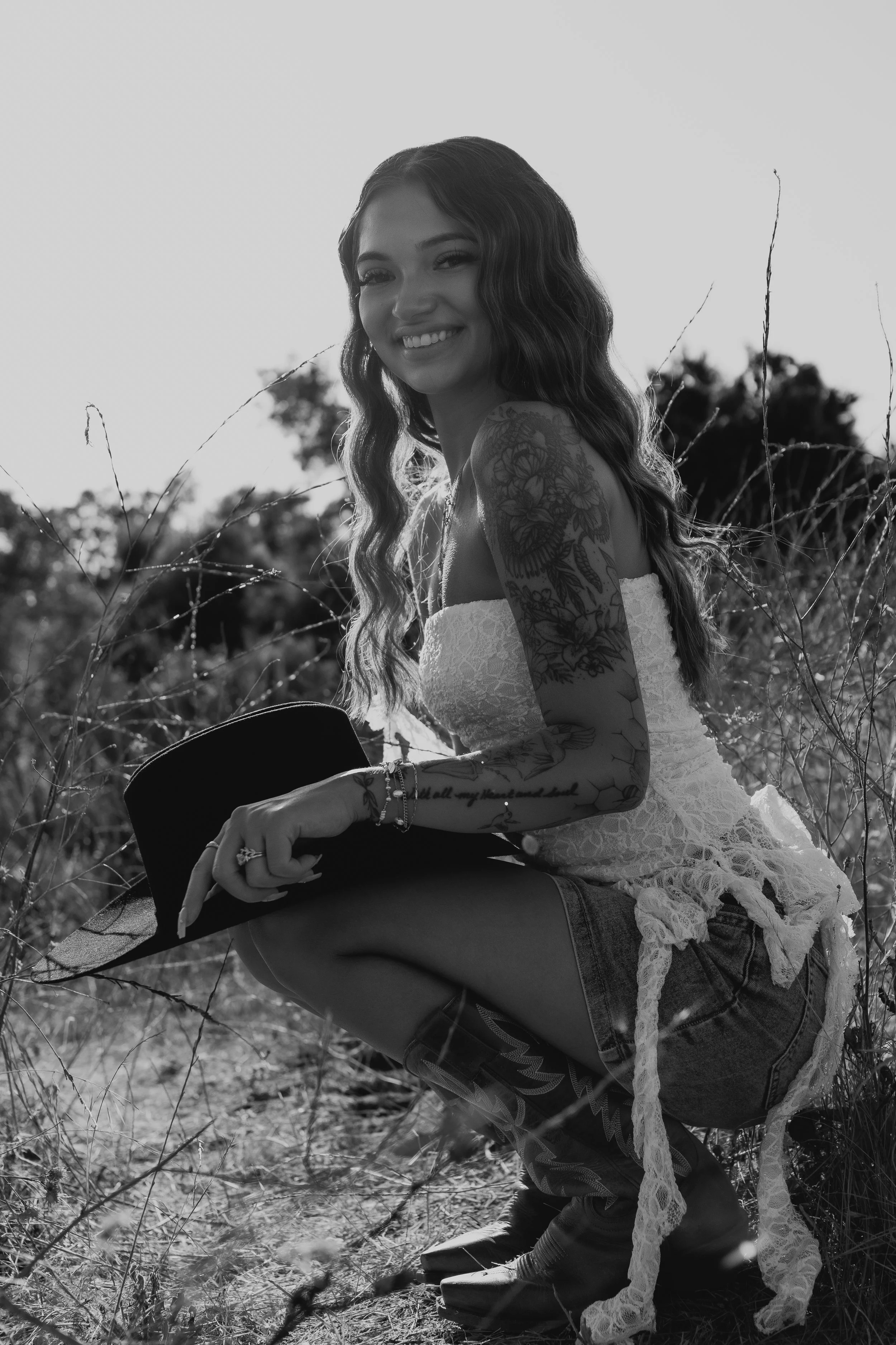 A smiling woman with long wavy hair and tattoos on her arm, wearing a strapless top and denim shorts, squatting outdoors among tall grass with trees in the background, holding a hat and a guitar.