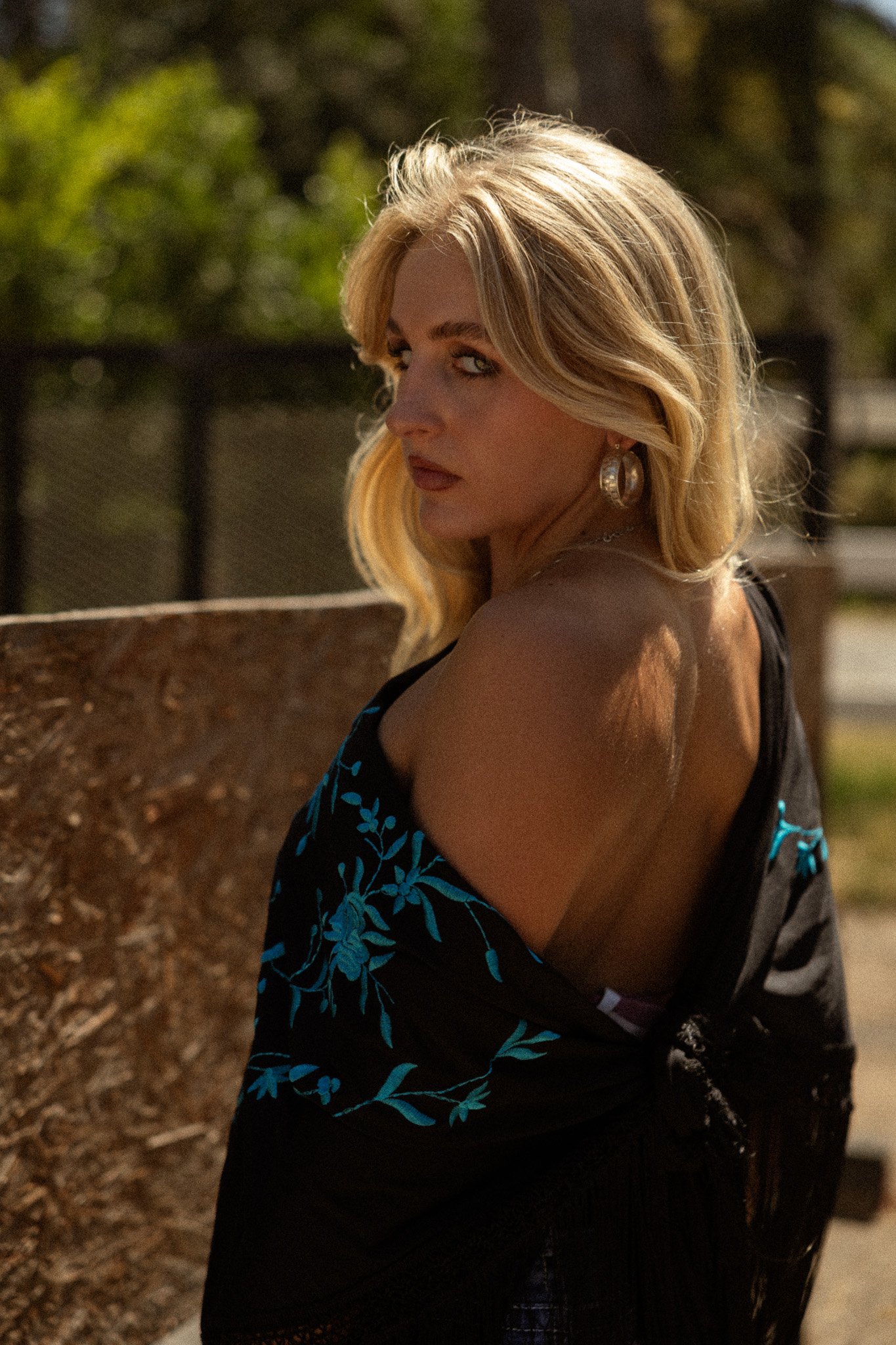 A blonde woman with wavy hair looking over her shoulder, wearing hoop earrings and a black top with blue floral embroidery, outdoors with trees and a black fence in the background.