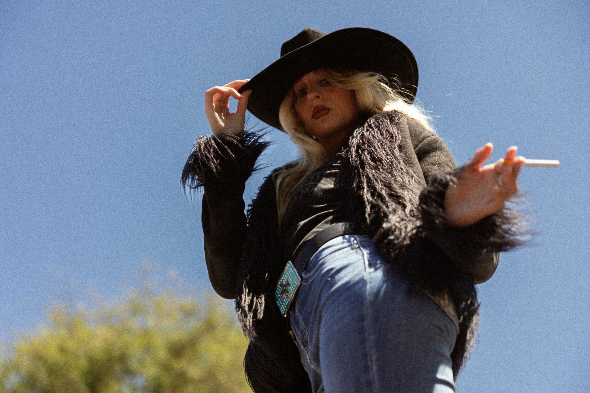 A woman with long blonde hair wearing a black wide-brimmed hat, a textured jacket, and blue jeans, holding a cigarette in her right hand, against a clear blue sky with some blurred greenery at the bottom.