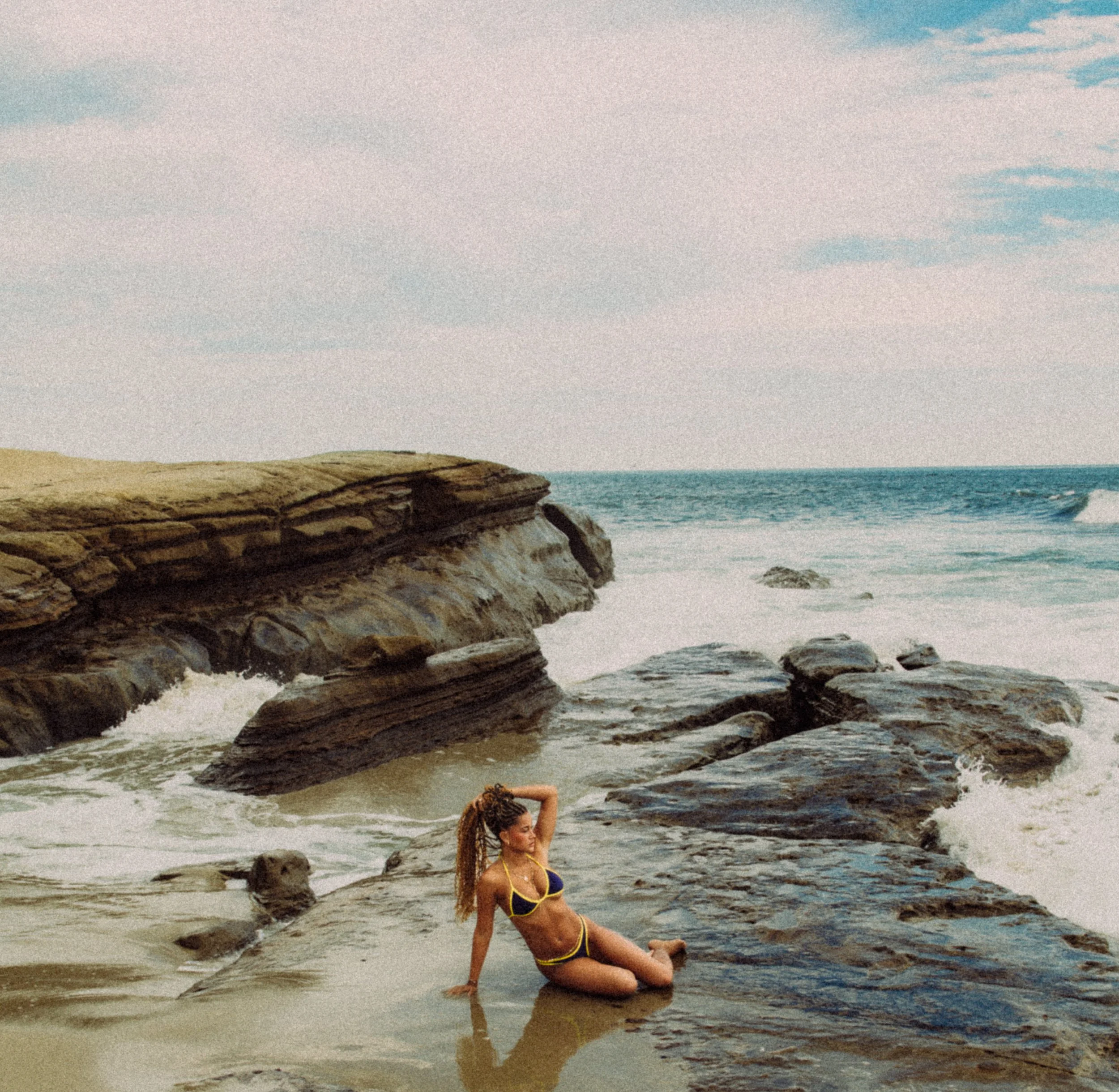 A woman in a bikini sitting on a rock on the beach, with the ocean and a cloudy sky in the background.