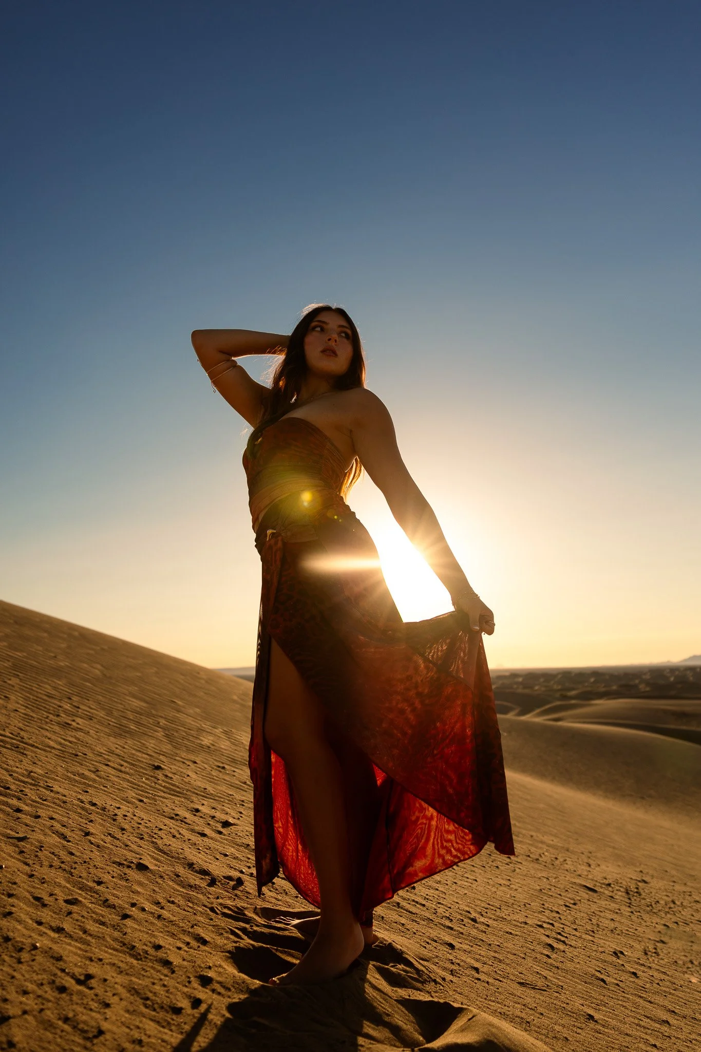Woman wearing a flowing red dress standing in a desert at sunset.