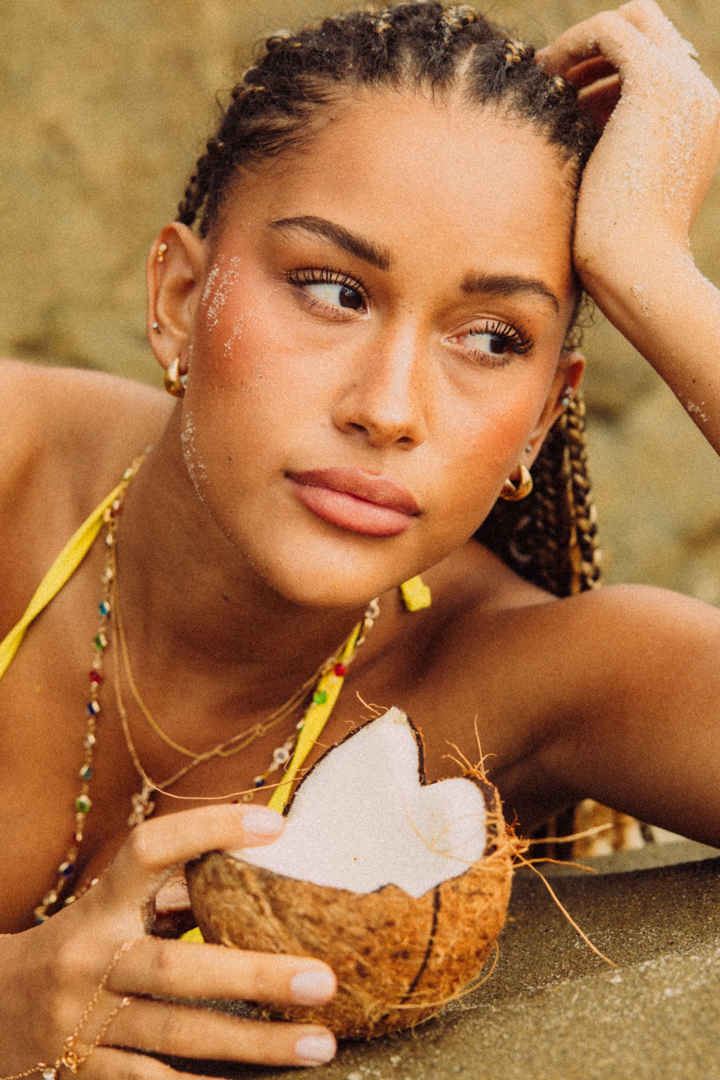 A woman with braided hair and jewelry is holding a broken coconut shell with a heart-shaped cutout, sitting outdoors with sand visible around.