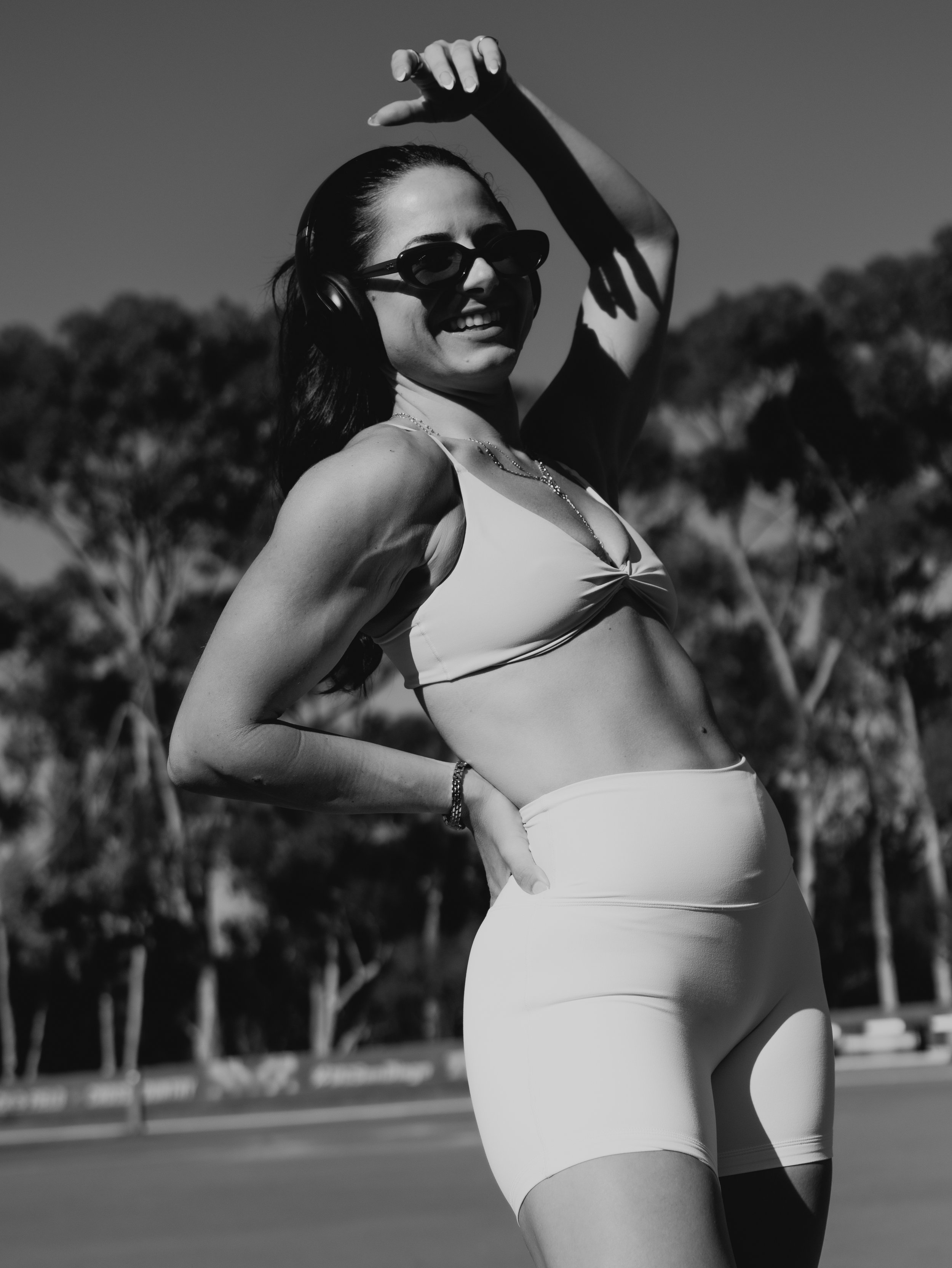 A woman wearing sunglasses and headphones, smiling, with her hand raised above her head, outdoors on a sunny day, with trees and a fence in the background.