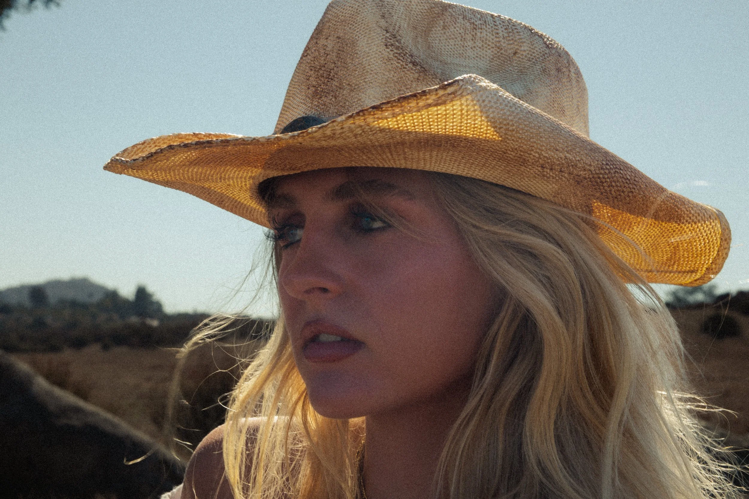 Close-up of a woman wearing a large, woven straw hat with a wide brim, outdoors on a sunny day, with blue sky and landscape in the background.