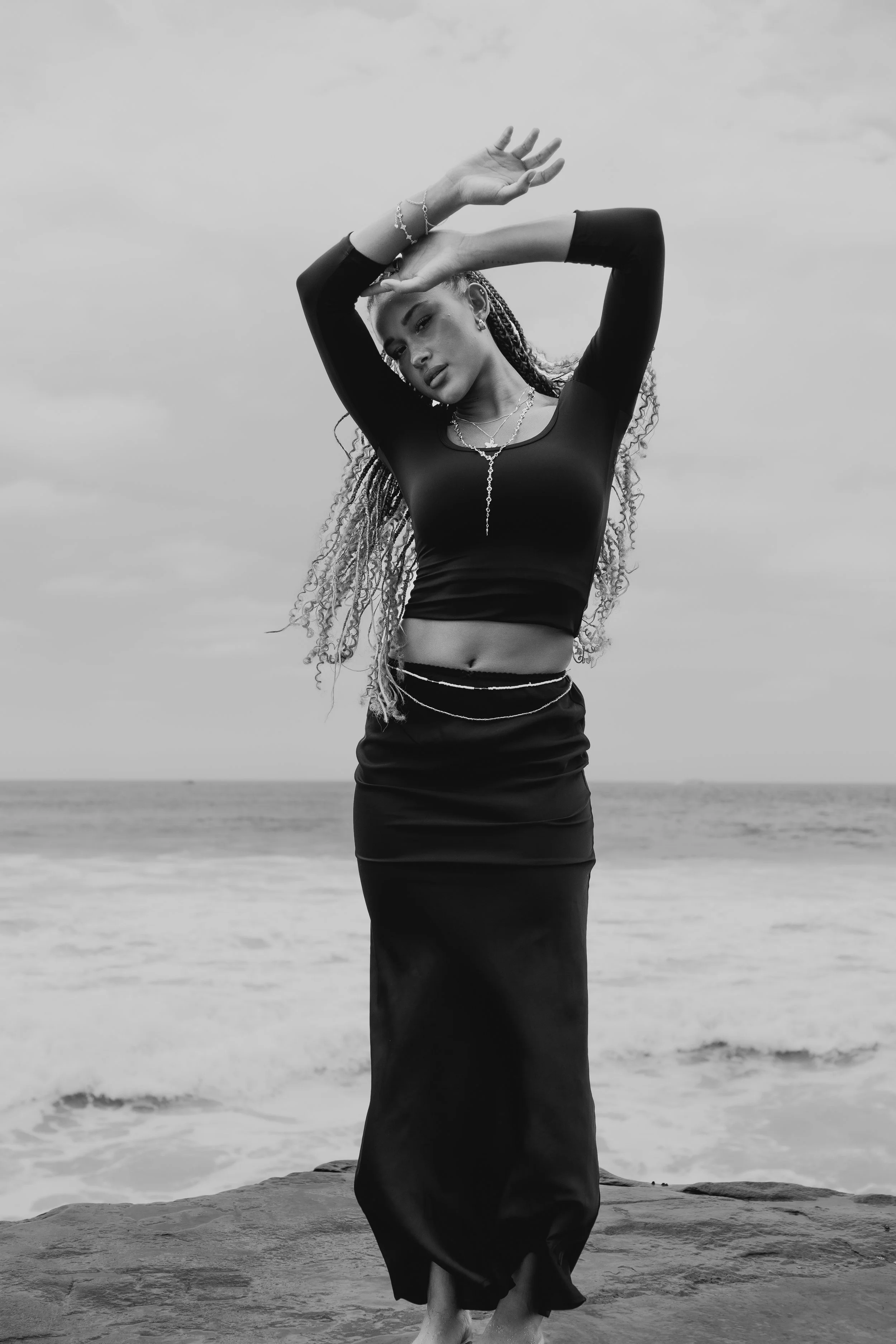 Black and white photo of a woman with long hair in braids, wearing a black long-sleeved crop top and a long black skirt, standing on rocks near the ocean with her arms raised above her head in a dance pose.