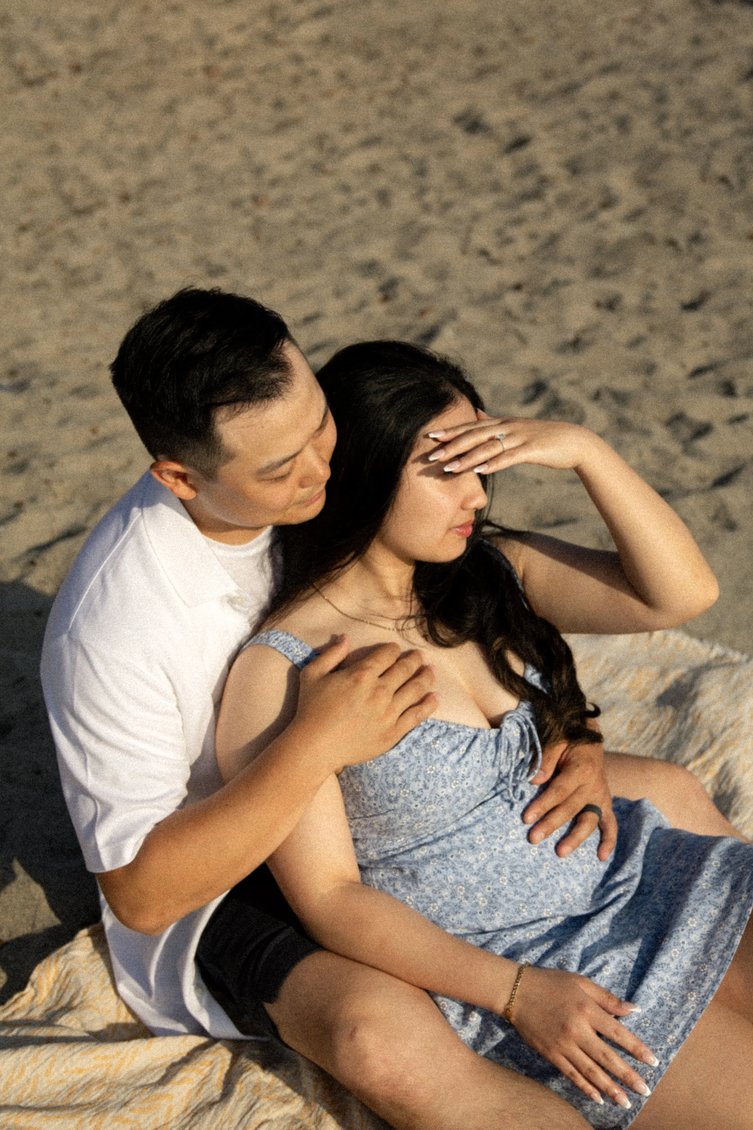 A man and woman sitting on a blanket on the sand at the beach. The man is hugging the woman from behind, and she is holding her hand on her forehead. They are close, intimate, and relaxed.