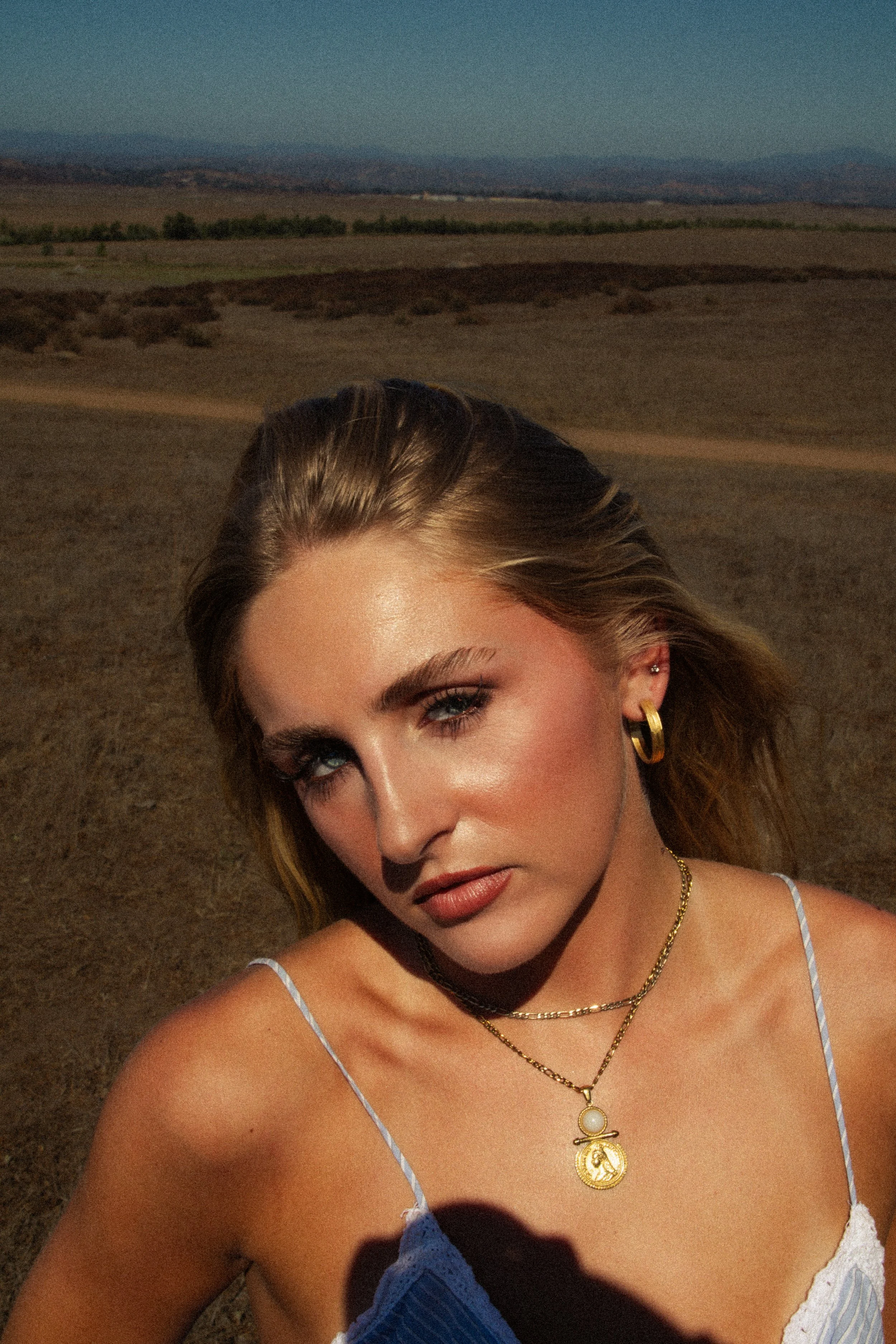 Close-up of a woman's face and shoulders, wearing layered gold necklace and earrings, with a blue and white dress, outdoors with a desert landscape, mountains, and clear sky in the background.