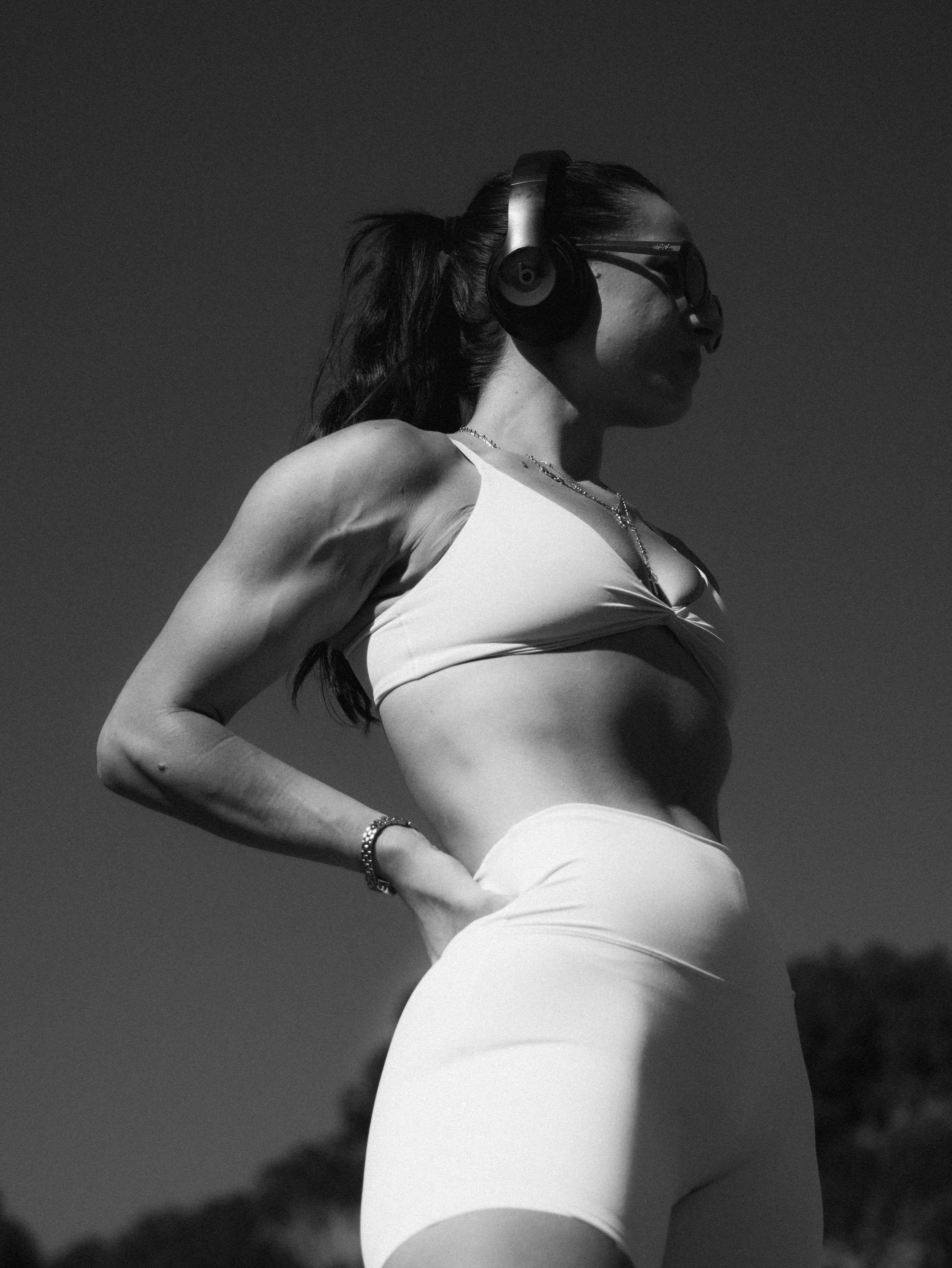 A woman with a ponytail wears headphones, glasses, and a tank top, standing outdoors with a dark sky in the background.