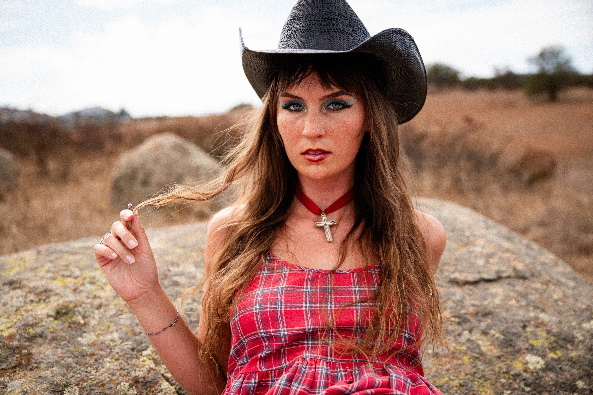 A woman with long wavy hair, wearing a black cowboy hat, red plaid dress, red choker necklace with a cross, and makeup with blue eyeshadow and red lipstick, sitting outdoors on a rocky landscape with trees in the background.