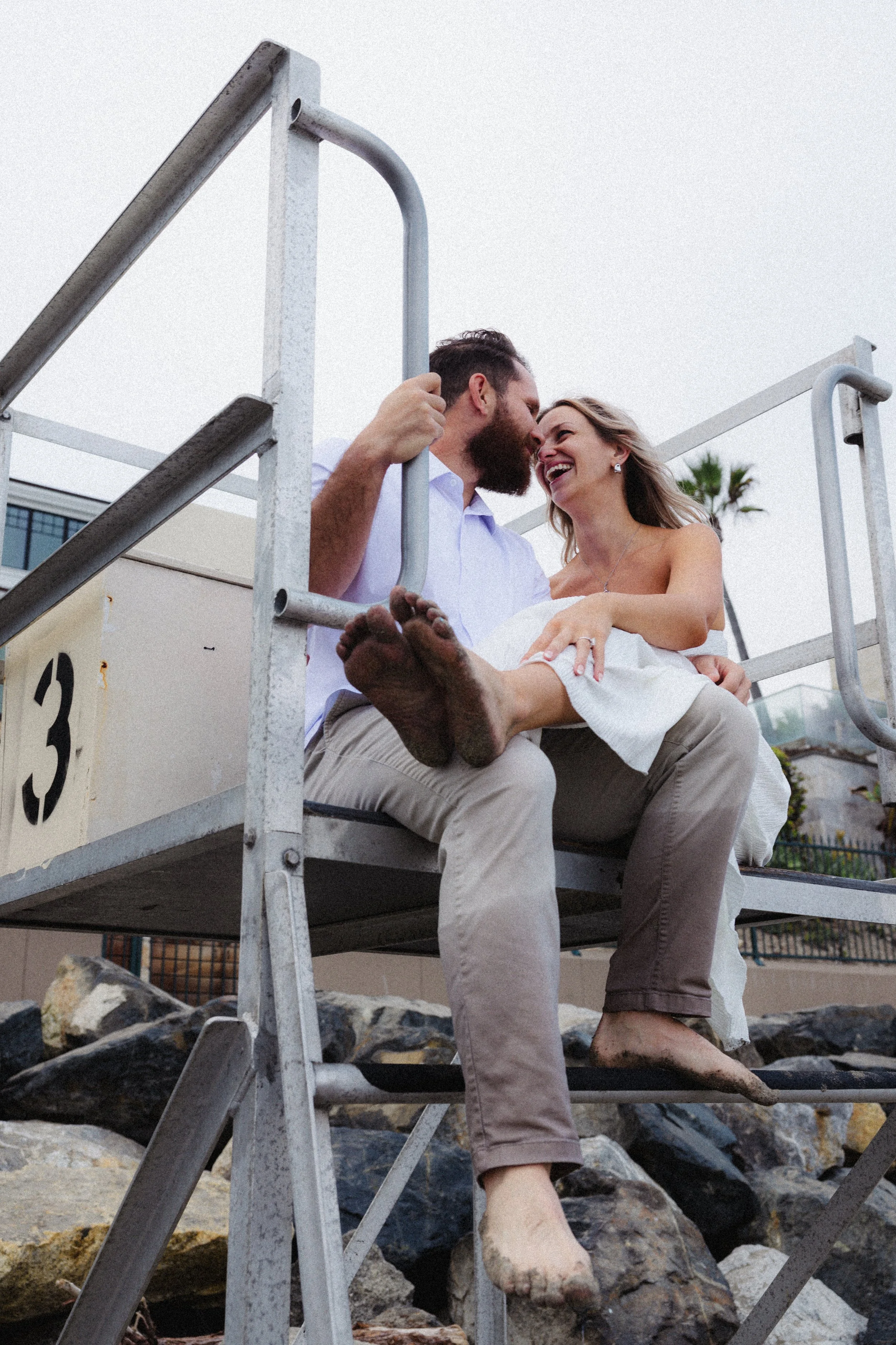 A couple sits closely together on a lifeguard stand, smiling and looking into each other's faces, with their feet touching. The woman appears to be pregnant, and they are outdoors near rocks and a tree, with a cloudy sky in the background.
