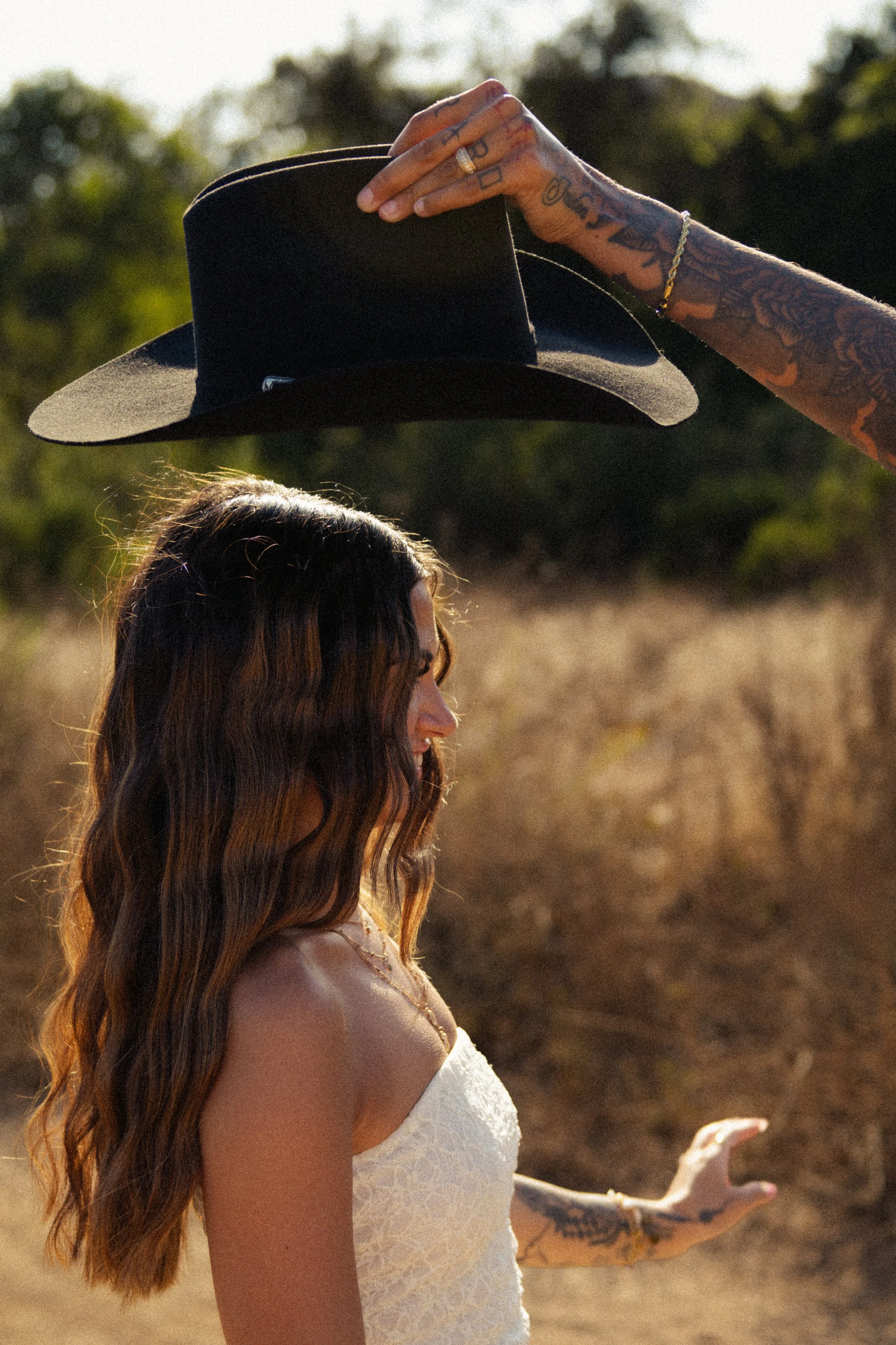 A woman with long, wavy hair and tattoos on her arm is holding a black hat in the air outdoors during daytime. The background shows a field and trees.