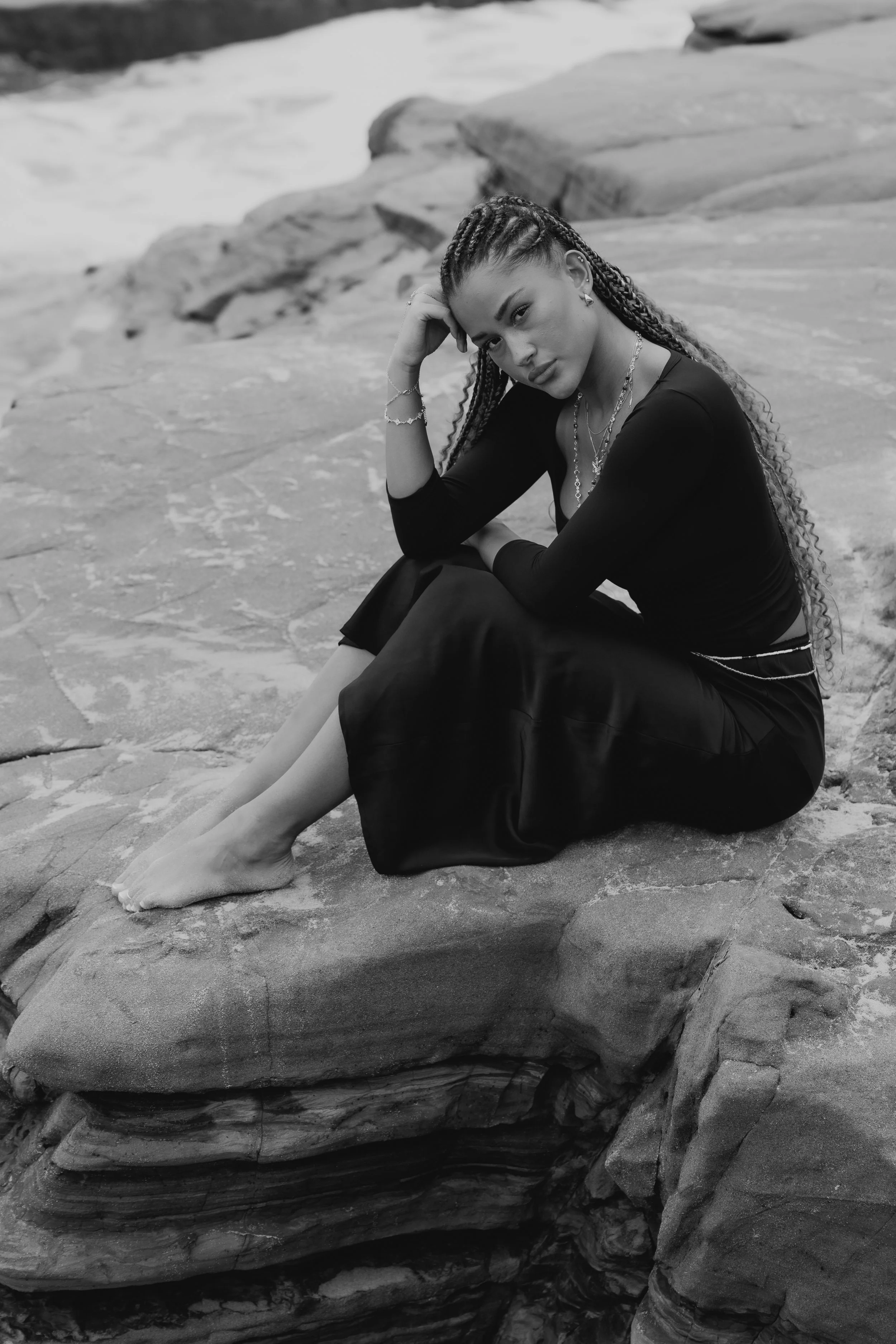 A woman with braided hair sits on rocks by the water, gazing at the camera. She wears jewelry and a black outfit, with her legs extended on the rocks.