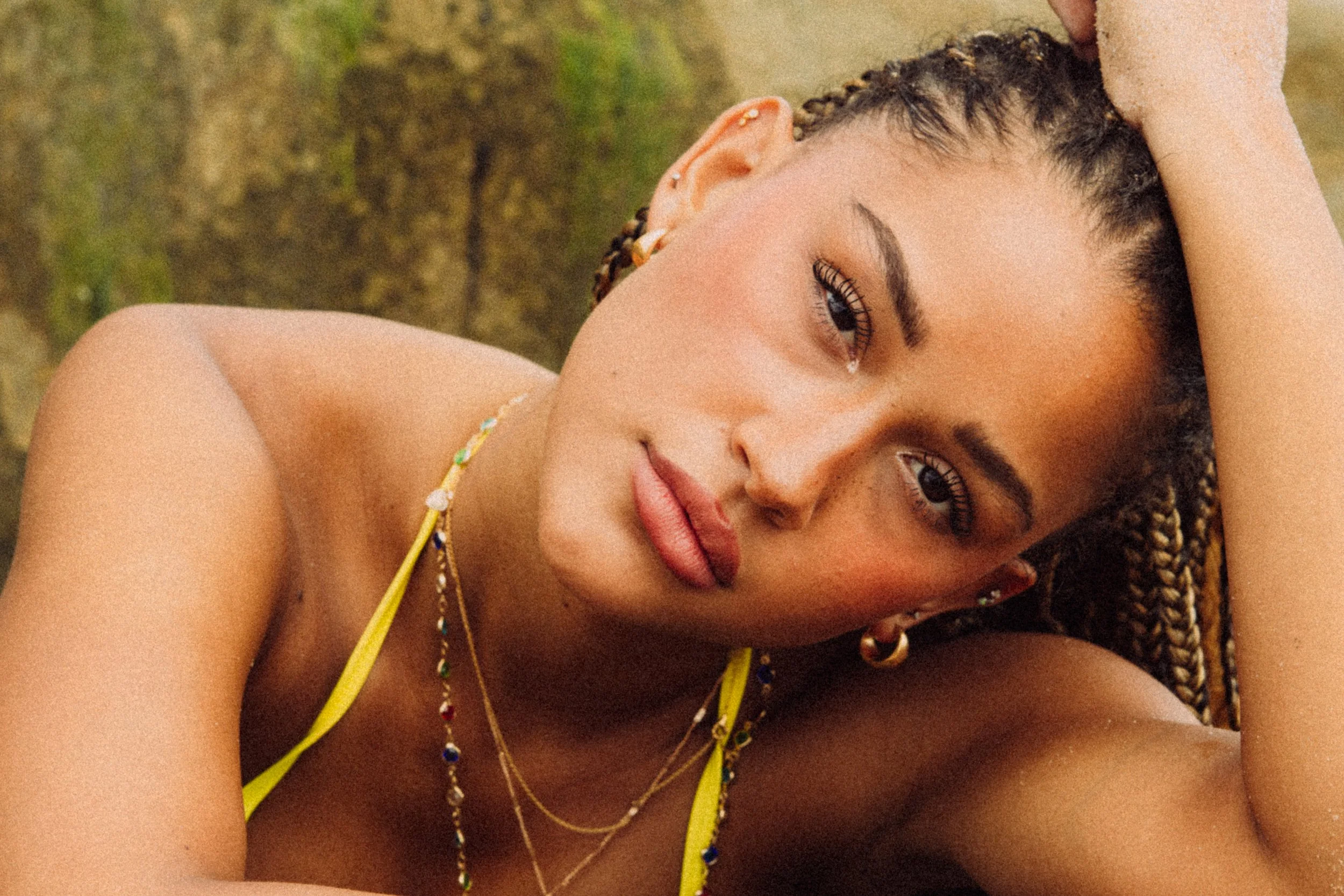 Close-up of a young woman with braided hair, wearing colorful jewelry and yellow clothing, lying down outdoors on grass.