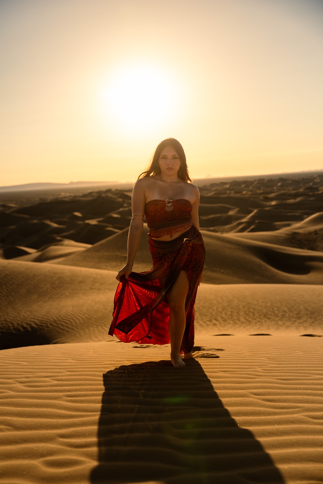 A woman wearing a red and black dress stands in a desert with sand dunes at sunset, casting a long shadow on the ground.